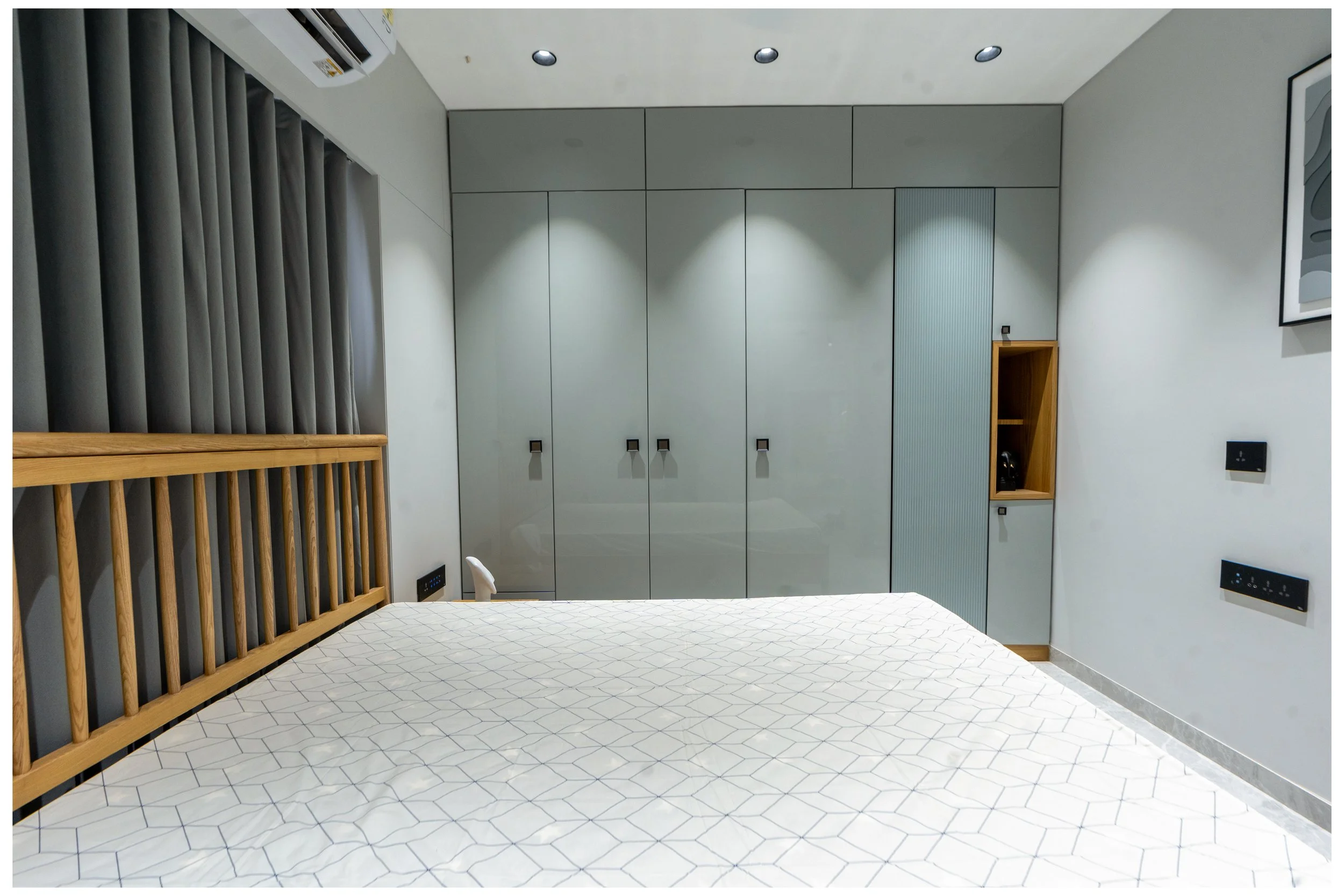 A modern bedroom with a wooden bed frame, white mattress, gray curtains, large gray closet, and small wall nook with a telephone. The room has a ceiling air conditioner unit and black electrical outlets.