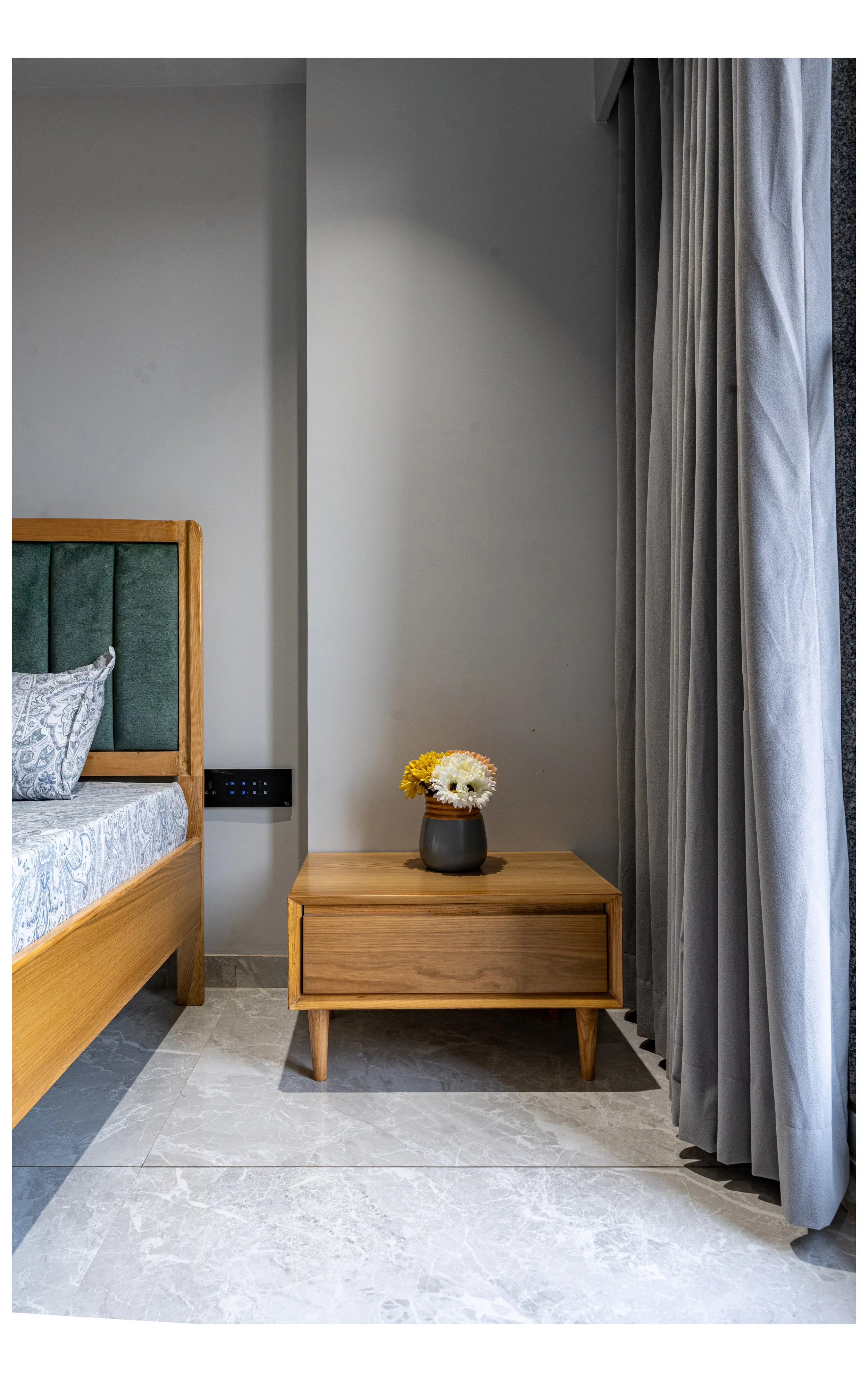 A modern bedroom with a wooden bed and an attached green upholstered headboard, adjacent to a wooden nightstand with a vase of yellow and white flowers, and floor-to-ceiling gray curtains next to a window.