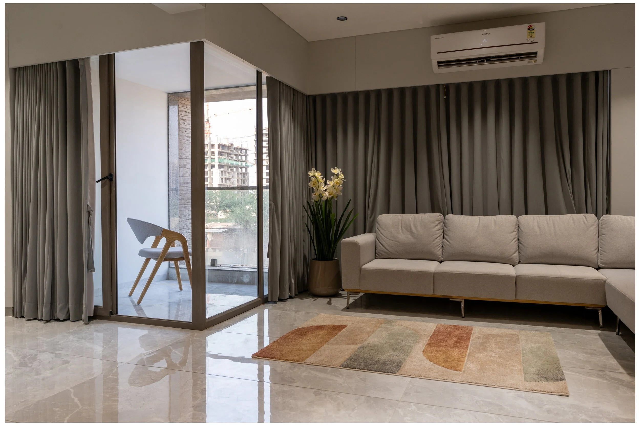 Modern living room with a beige sofa, a potted plant with white flowers, a colorful area rug, and a glass door leading to a balcony with an outdoor chair. Gray curtains hang on the windows, and an air conditioning unit is mounted on the wall.