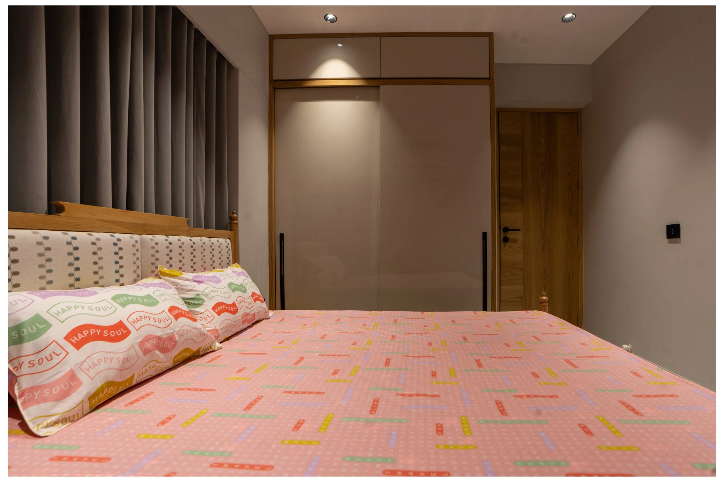 A bedroom with a bed, patterned pillowcases, and a pink bedsheet, alongside a wooden wardrobe with beige sliding doors and a door on the right side.