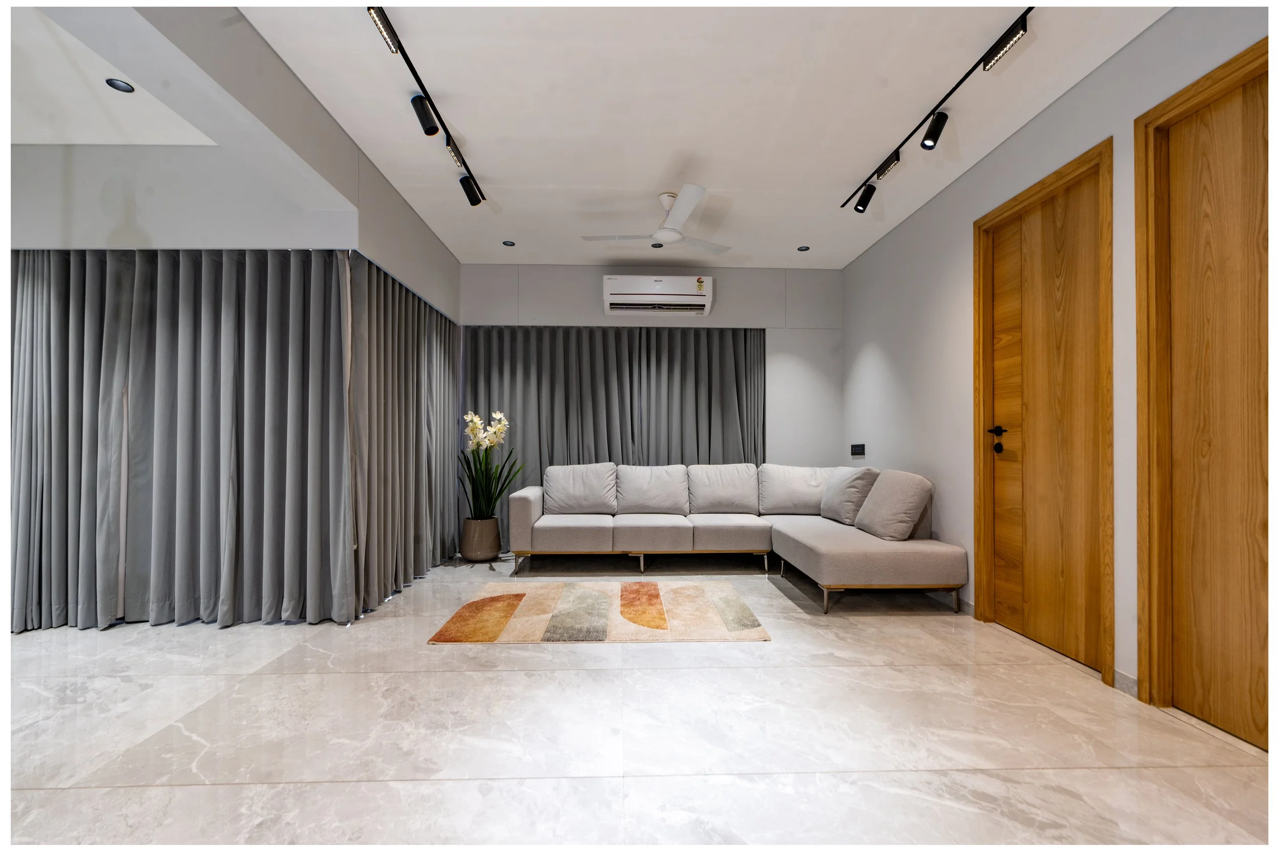 Modern living room with a white sectional sofa, potted plant with white flowers, gray curtains, wooden doors, and track lighting.