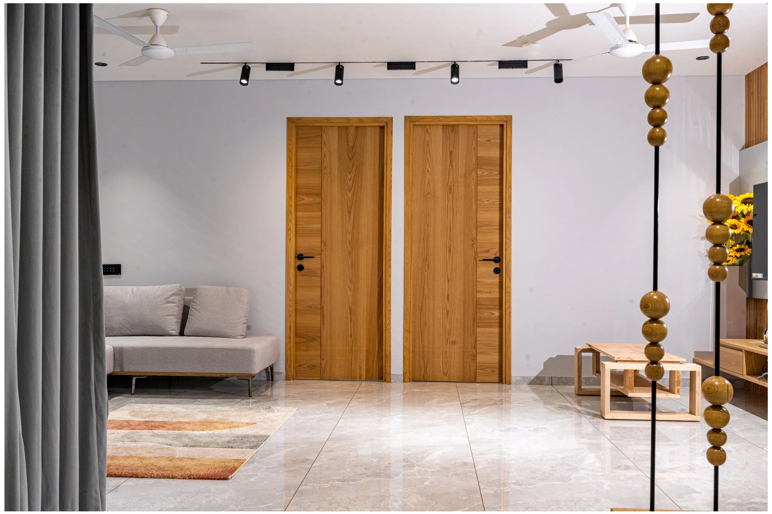 Interior view of a modern living room with two wooden doors, a gray sofa, a colorful striped rug, and decorative elements including a beaded floor divider, ceiling fans, and sunflowers on a shelf.