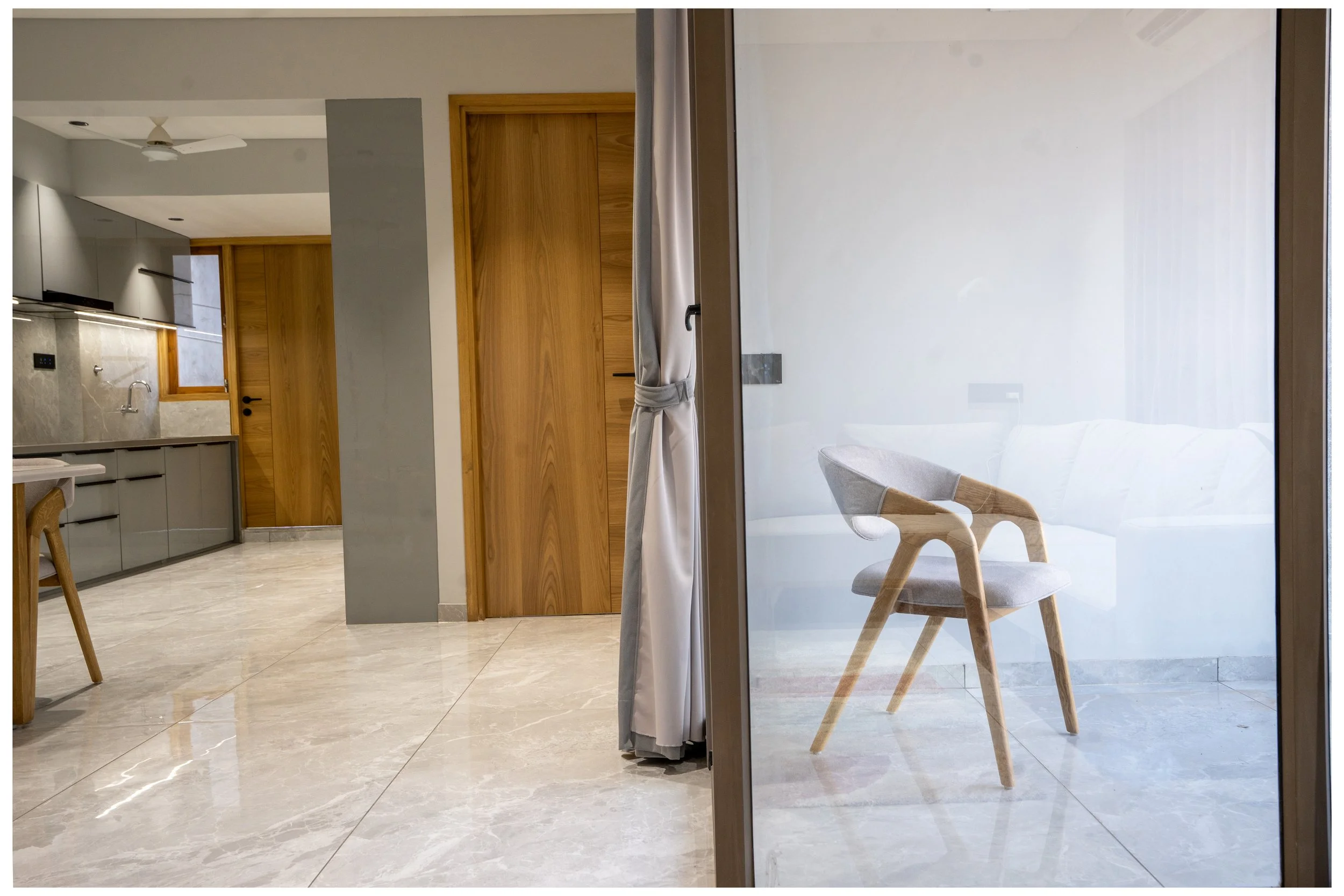 Interior view of a modern living space, showing part of a kitchen with gray cabinets and a wooden door, a sitting area with a chair and a sofa visible through a glass partition with a curtain.