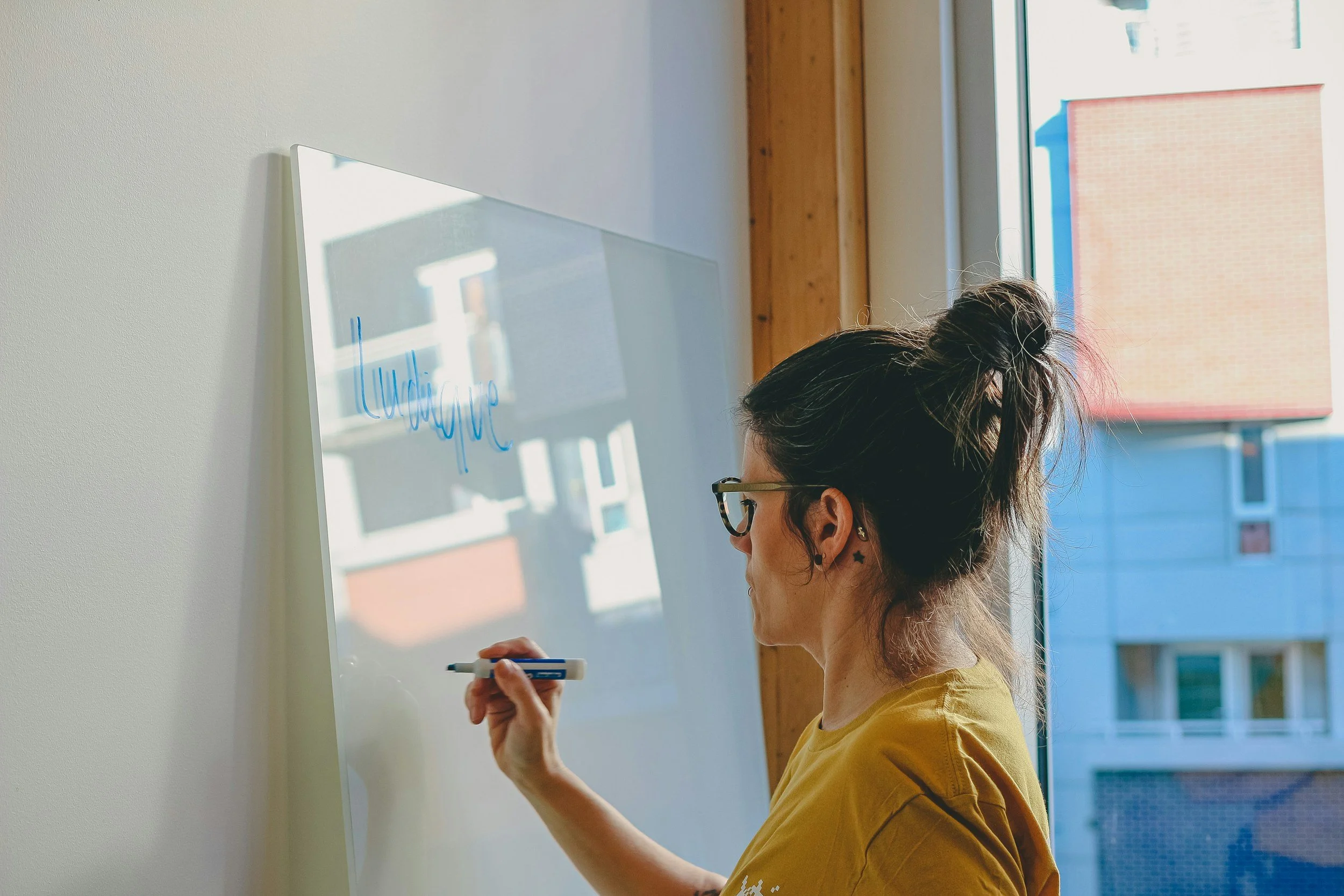 A woman with dark hair tied in a messy bun, wearing glasses and a yellow shirt, writes on a transparent whiteboard with a marker in front of a window. Everyday EA.