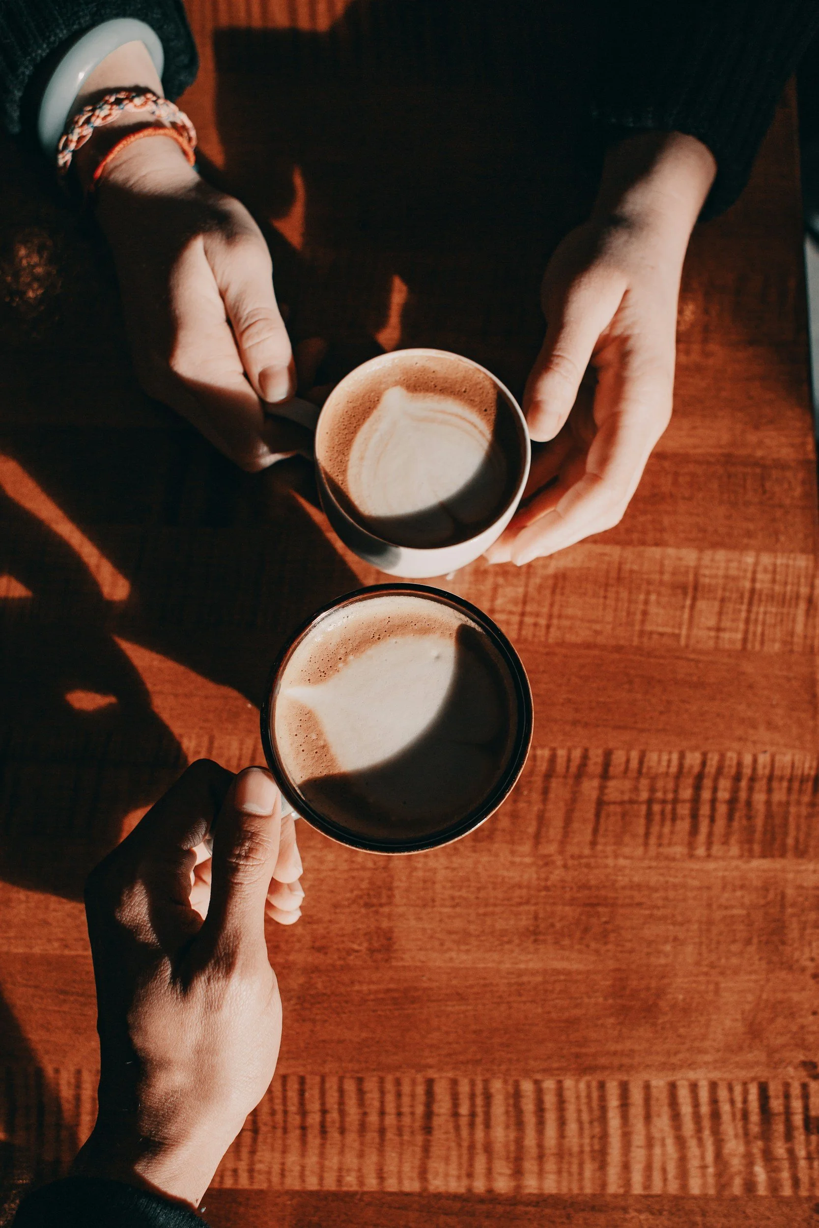 Two people are clinking mugs filled with coffee, viewed from above on a wooden table, with sunlight casting shadows. Everyday EA.