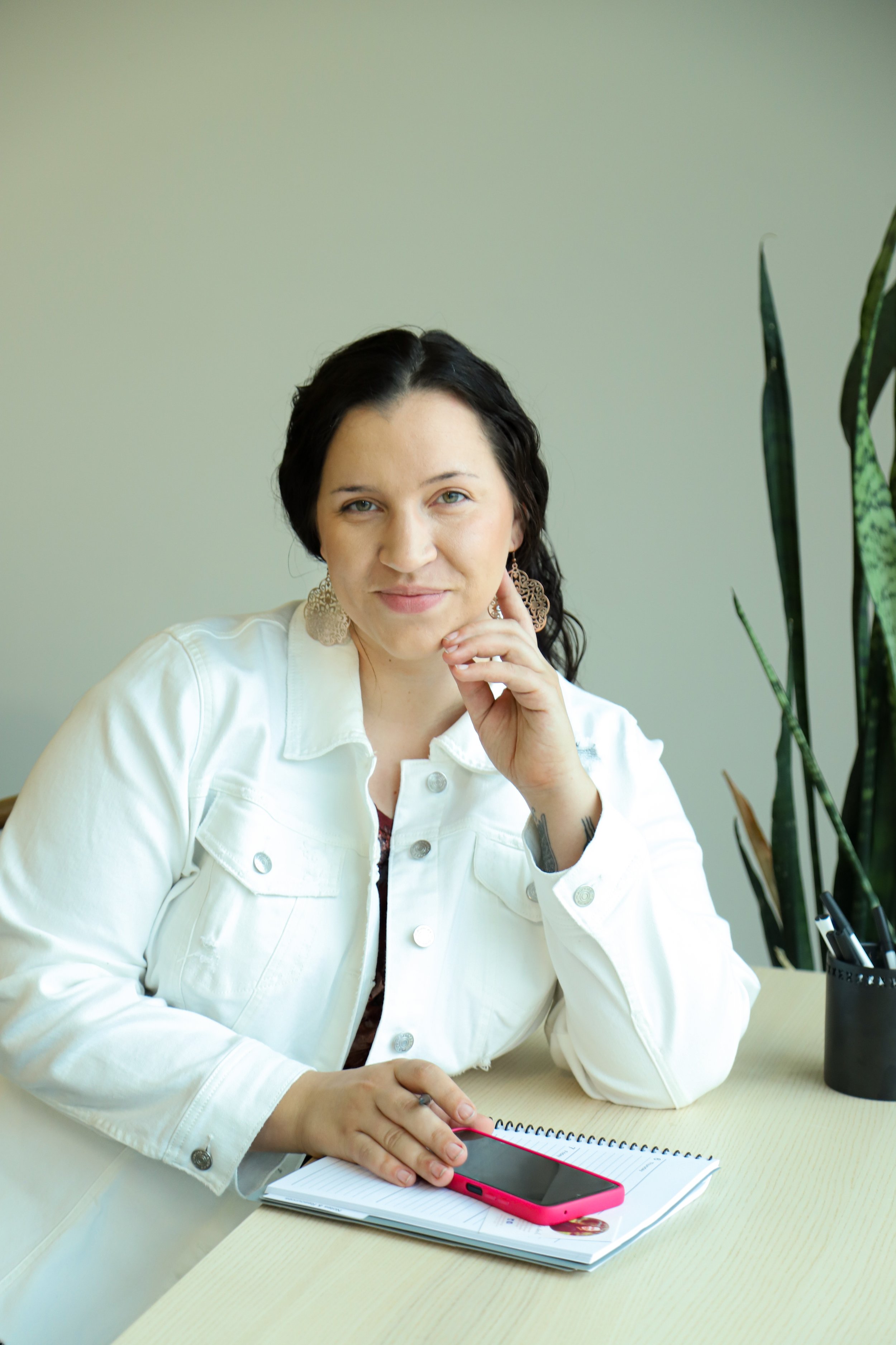 A woman with dark brown hair, dressed in a white jacket, sitting at a light-colored desk with a notebook and a pink smartphone, smiling slightly at the camera. Everyday EA.