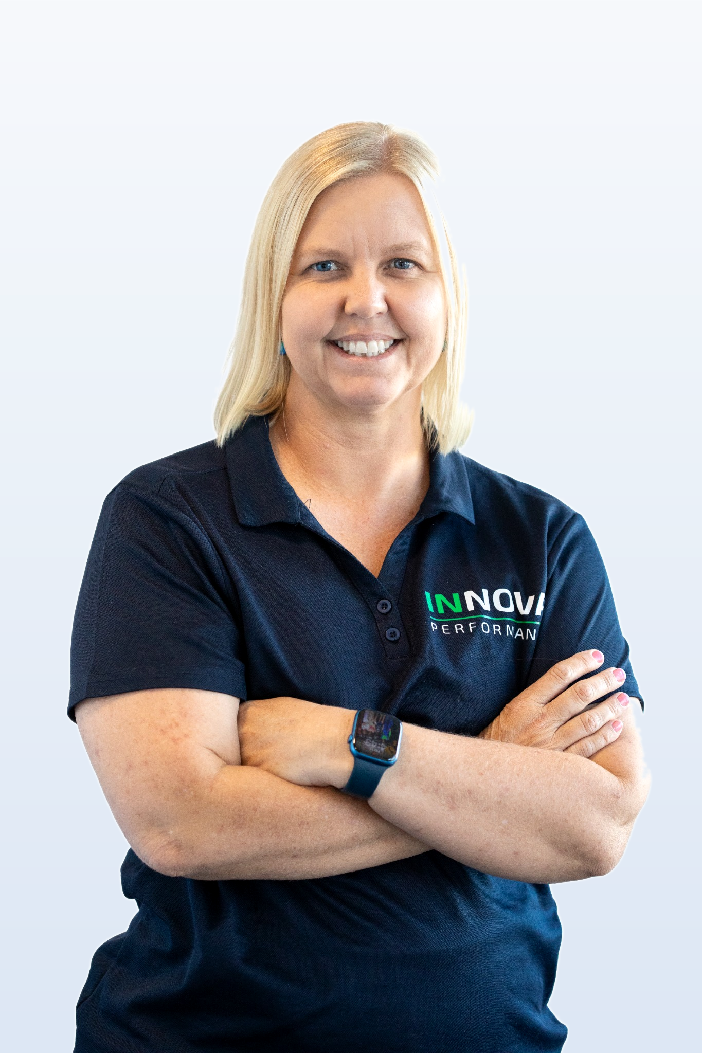 A woman with blonde hair, wearing a navy blue polo shirt with 'INNOVA PERFORMANCE' logo, and a smartwatch, smiling with arms crossed against a light background.