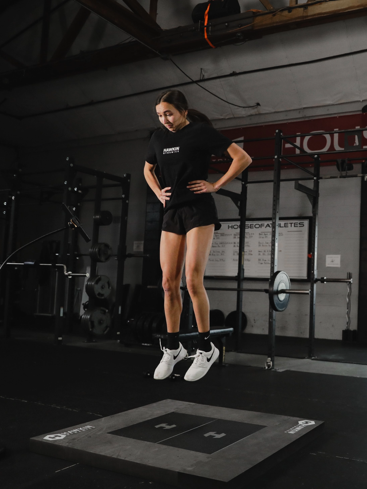 A woman wearing a black T-shirt, black shorts, and white Nike sneakers is jumping on a boxing training mat in a gym.