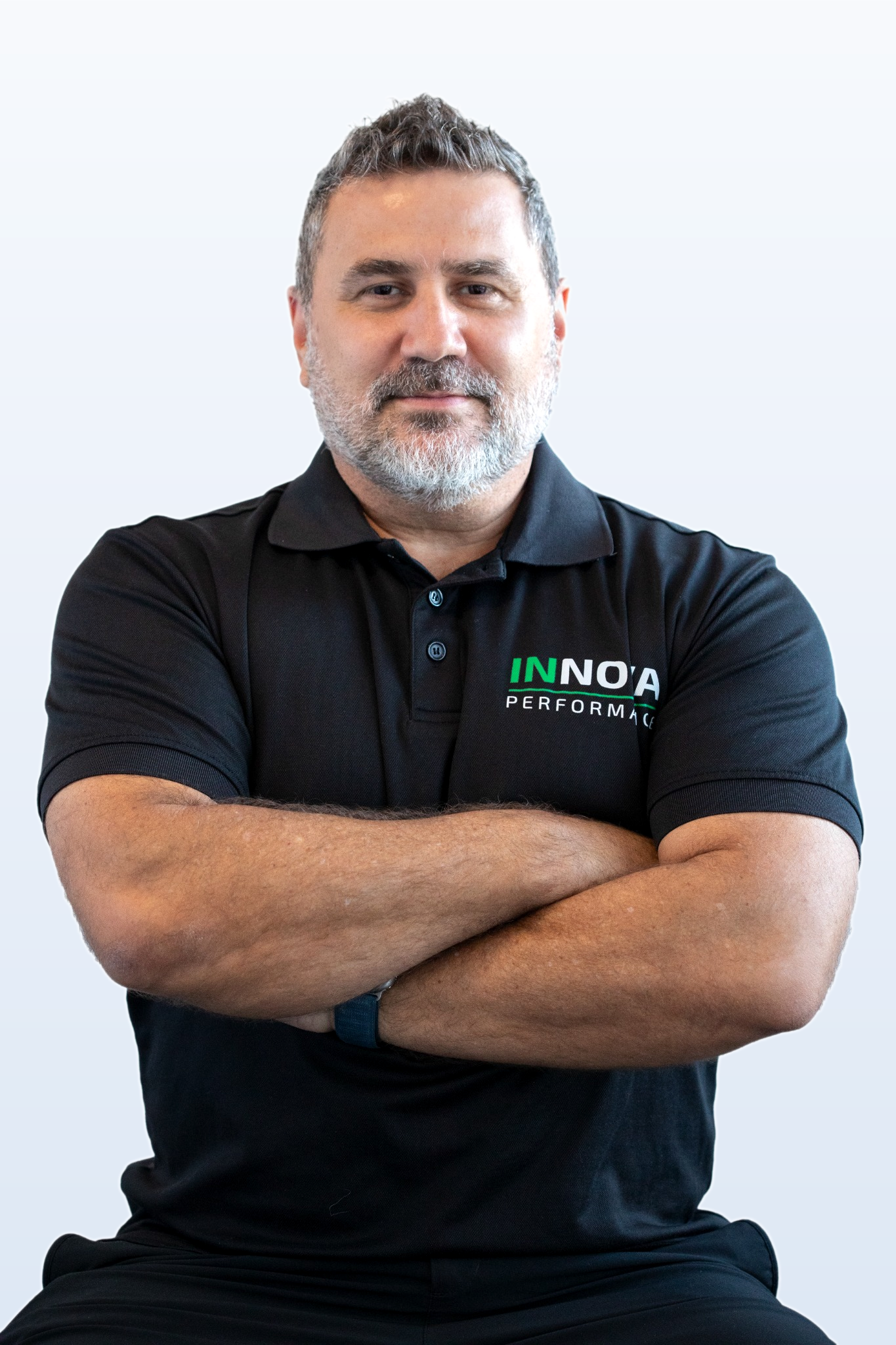 A middle-aged man with gray hair and beard, crossing his arms and smiling, wearing a black polo shirt with the logo 'INNOA Performance'.