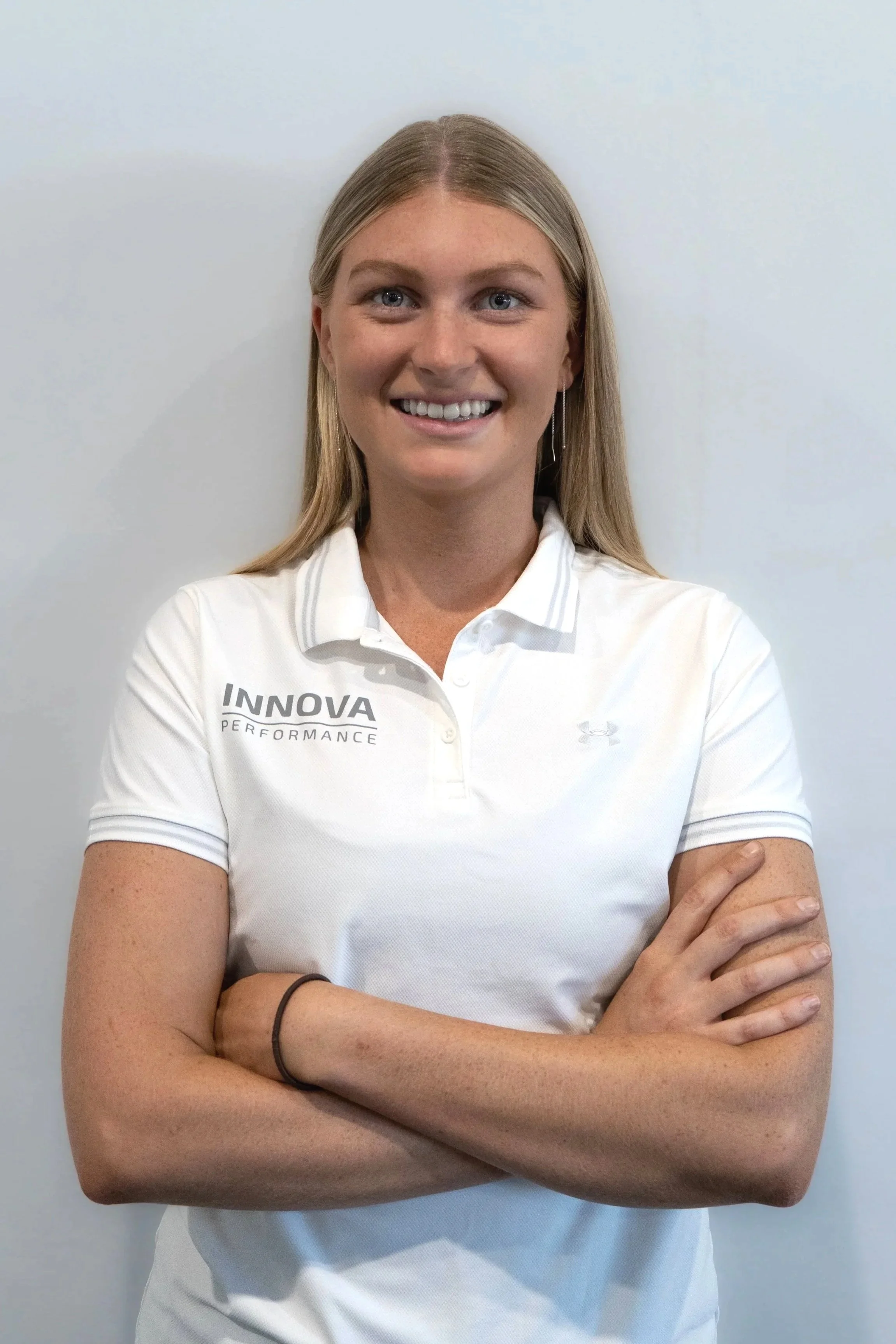 A woman with long blonde hair, smiling and crossing her arms, wearing a white polo shirt with 'INNOVA PERFORMANCE' embroidered on it, standing against a plain white wall.