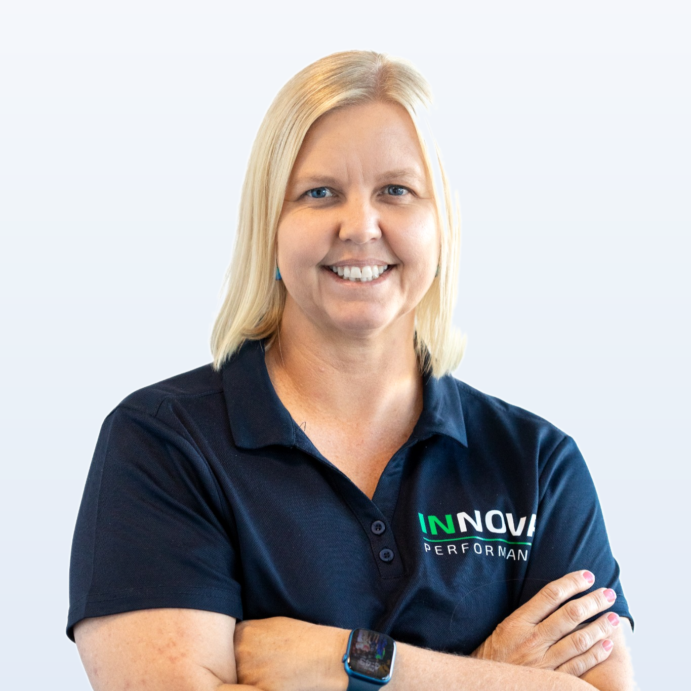 A smiling blonde woman wearing a navy blue polo shirt with 'INNOVA Performance' logo, standing with arms crossed against a light background.