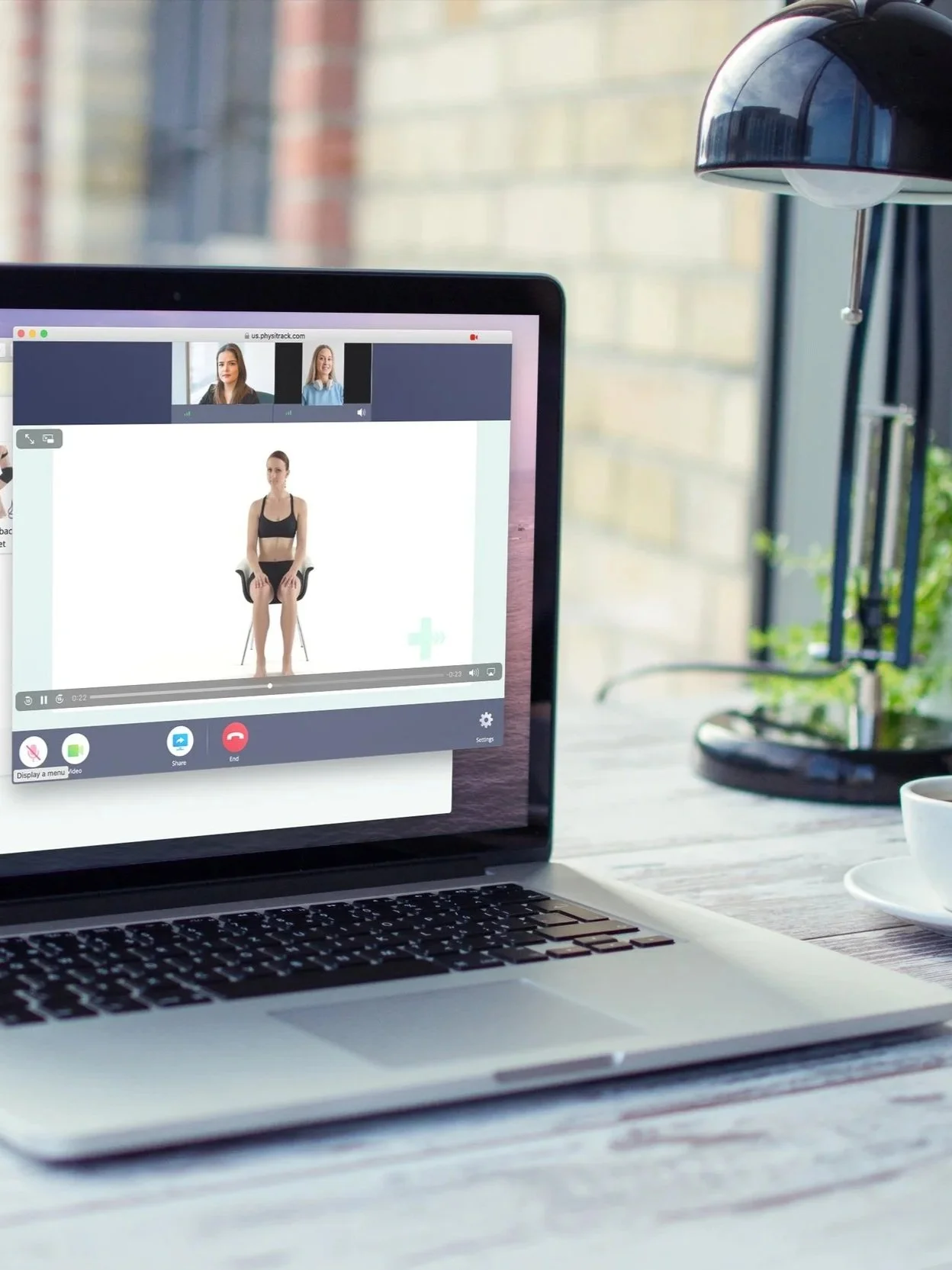 Laptop on a wooden table showing an online video call with a woman sitting on a chair, and two other women in smaller windows at the top. There is a white cup and saucer, a desk lamp, and a potted plant next to the laptop.