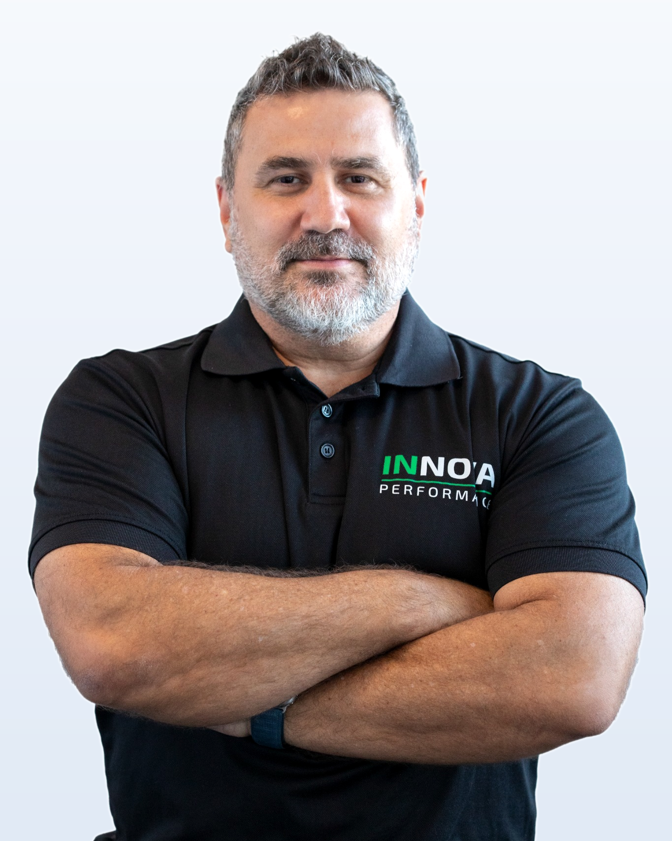 A man with short gray hair and a beard, wearing a black polo shirt with 'INNOVA PERFORMANCE' logo, standing with arms crossed against a plain light gray background.