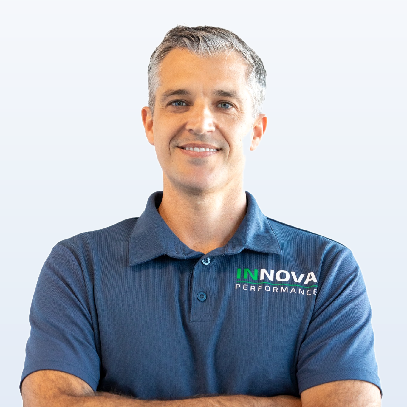 Portrait of a man with gray hair wearing a navy blue polo shirt with 'Innova Performance' logo, smiling with arms crossed against light background.