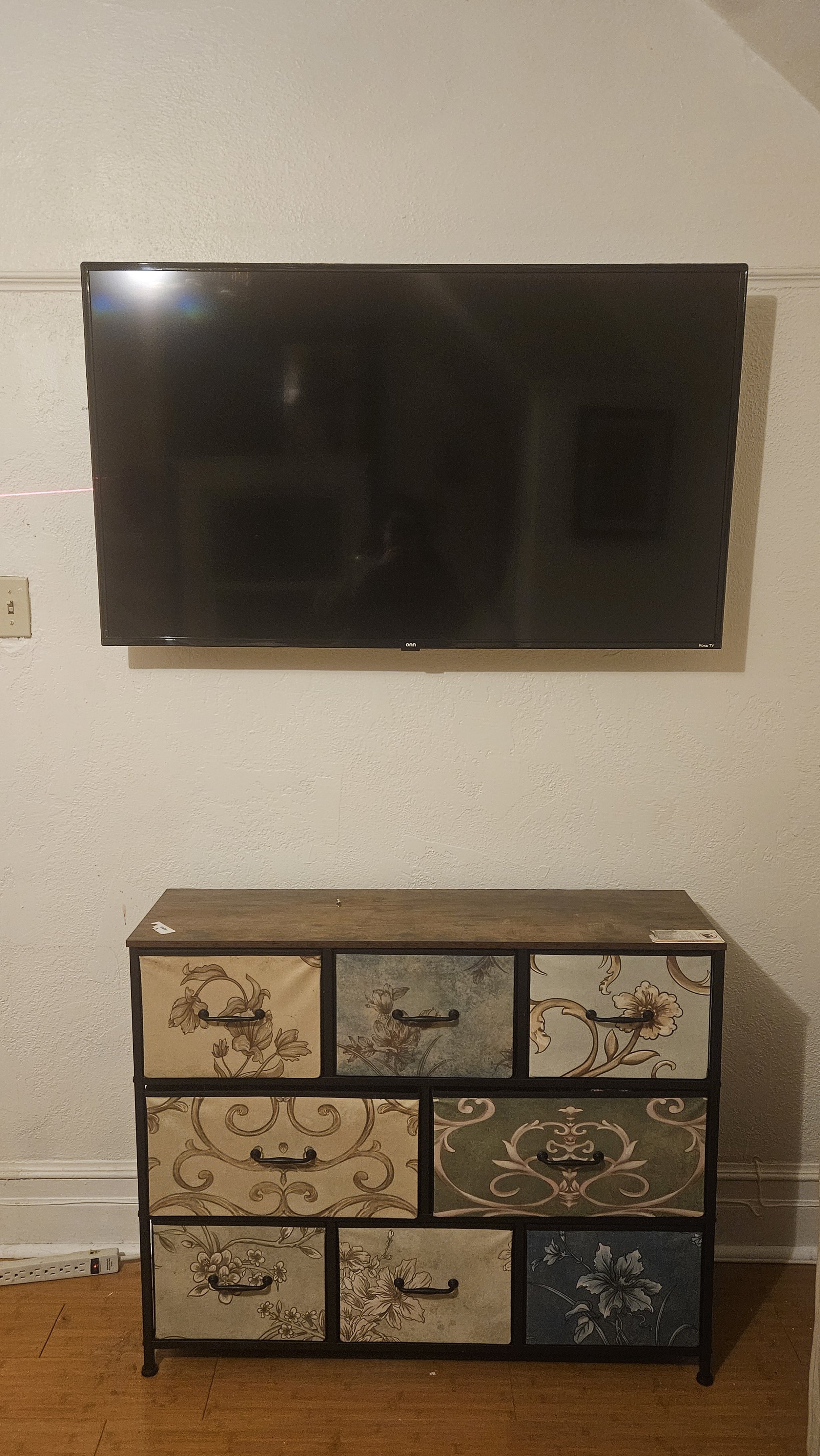 Wall-mounted flat-screen television above a vintage-style dresser with floral patterns and black handles in a room with white textured walls and wooden floor.