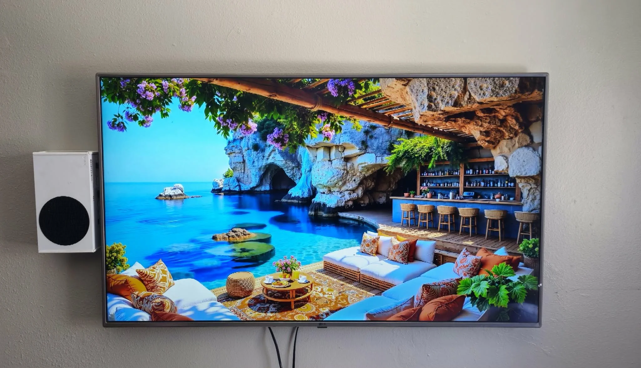 Entertainment room featuring a large mounted flat-screen TV displaying an outdoor seaside lounge, with a bar, colorful cushions, and greenery.