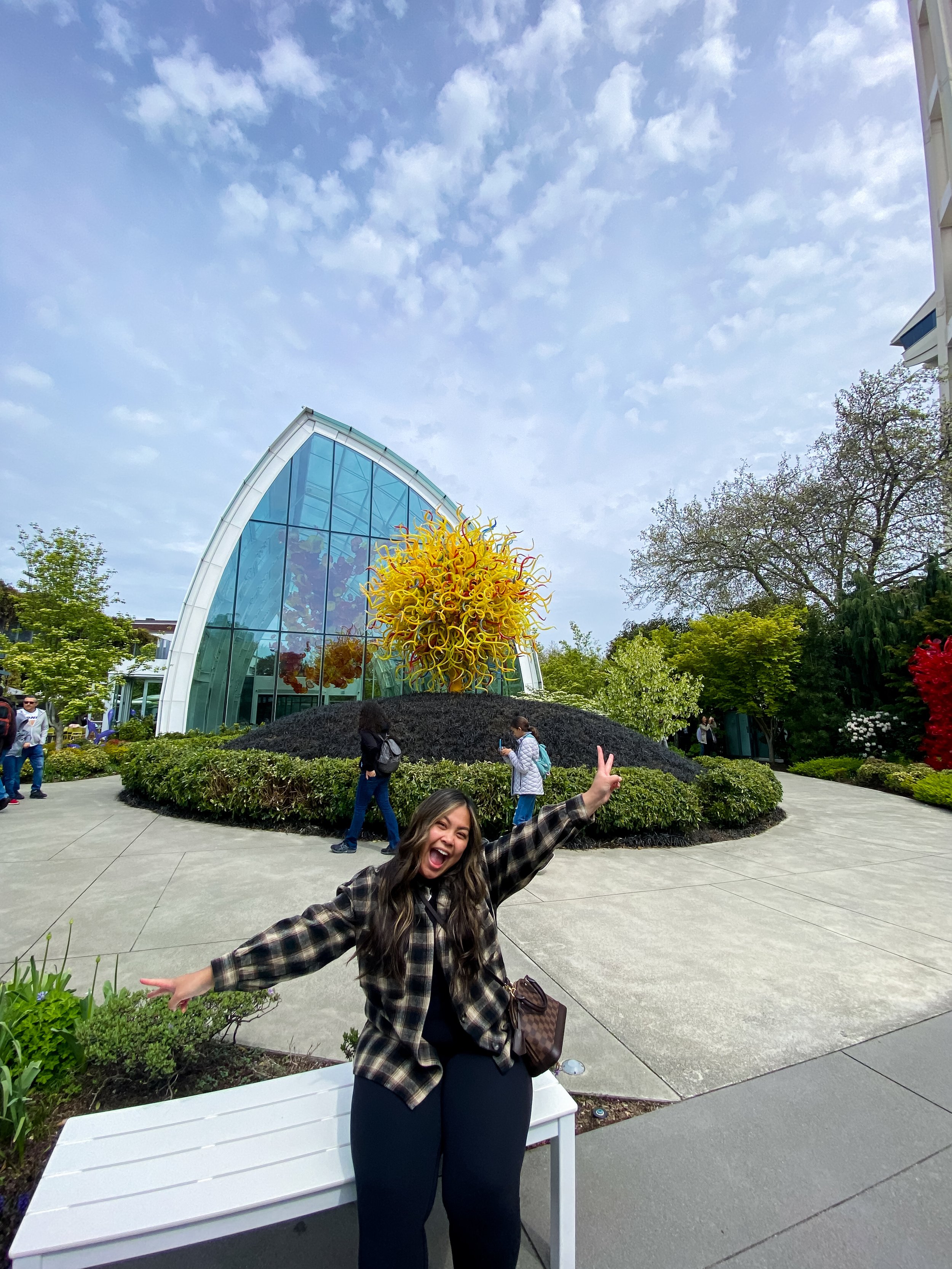 Young woman smiling and making a peace sign with her hand in front of a glass art installation shaped like a flower at a botanical garden.