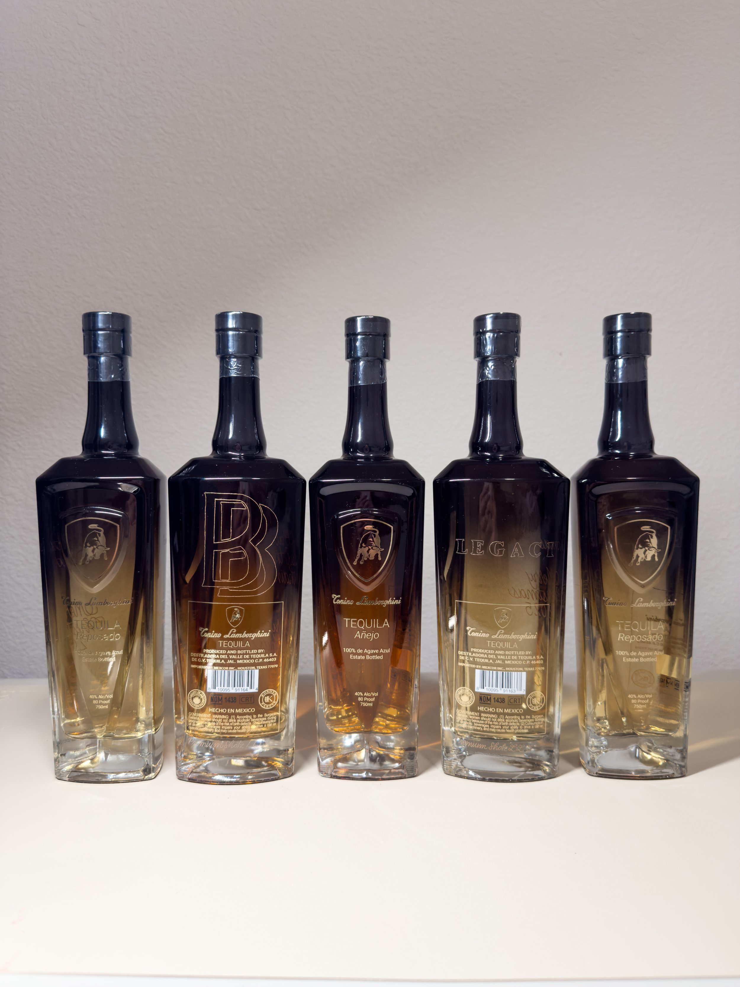 Five bottles of tequila lined up on a light-colored surface against a plain wall, with one bottle in the middle containing a darker liquid.