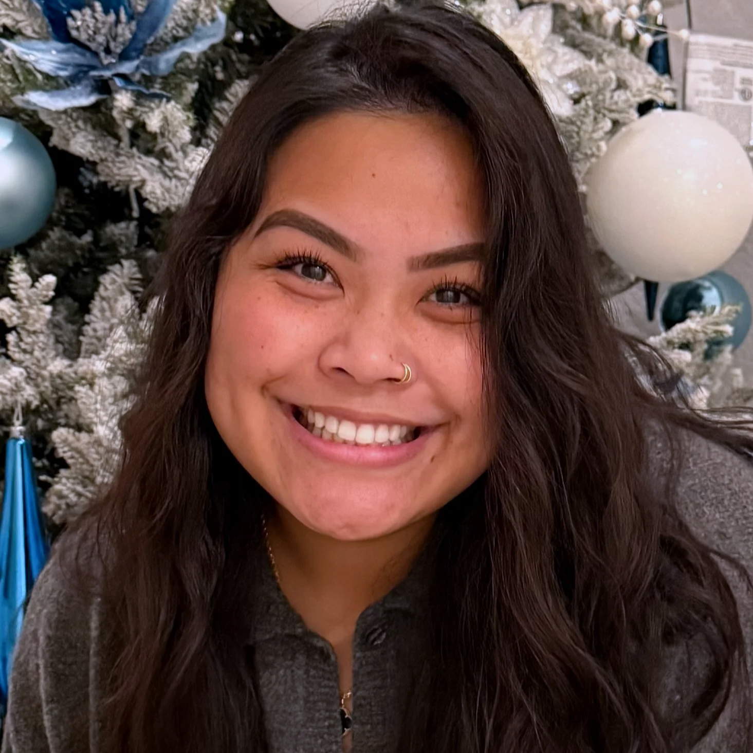 A smiling woman with long dark hair, wearing a nose ring, in front of a decorated Christmas tree with white and blue ornaments.