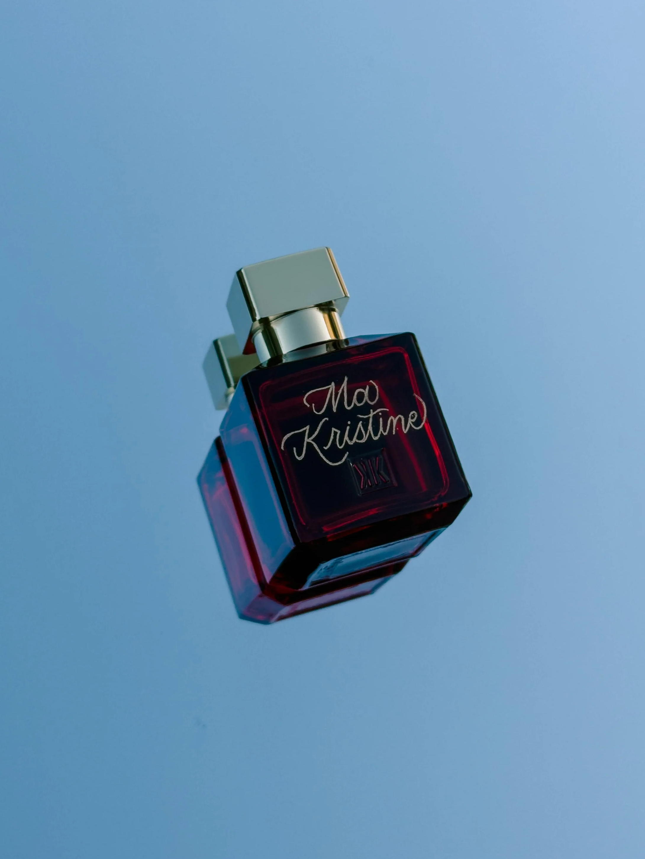A red perfume bottle with silver cap and black writing that says "Maa Kristine" on a light blue background.