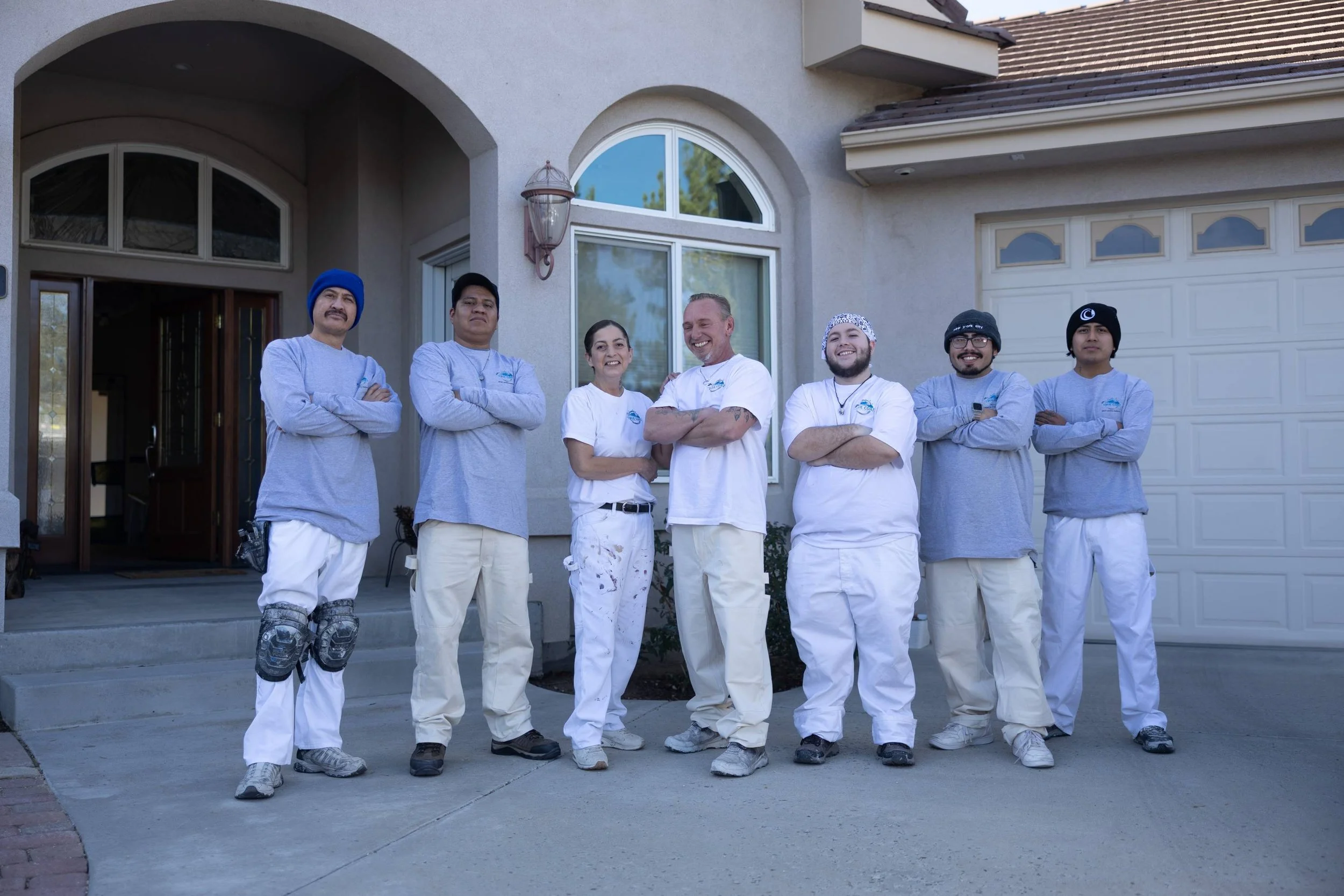 Group of seven maintenance or repair workers standing in front of a house, smiling, with arms crossed, dressed in work shirts and pants, some with hats or beanies, and some with work gloves or protective gear.