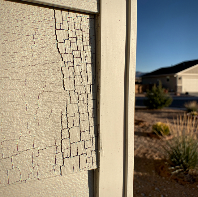 Cracked brick wall with white trim next to desert landscape with distant houses under blue sky.