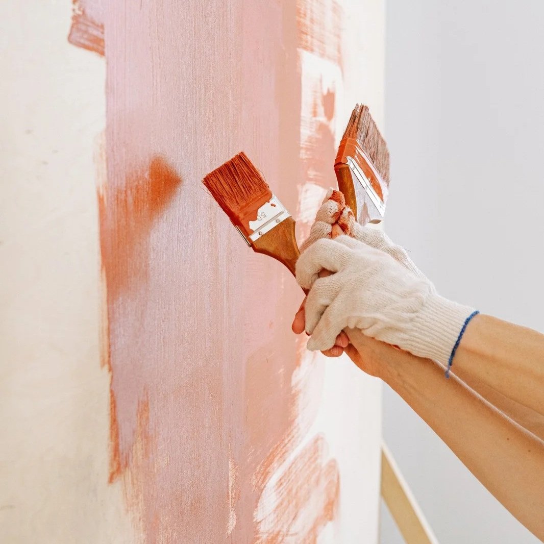 Close-up of gloved hands painting an interior wall with brushes. Fresh paint is being applied evenly, illustrating home repainting, wall refinishing, and professional interior decorating services.