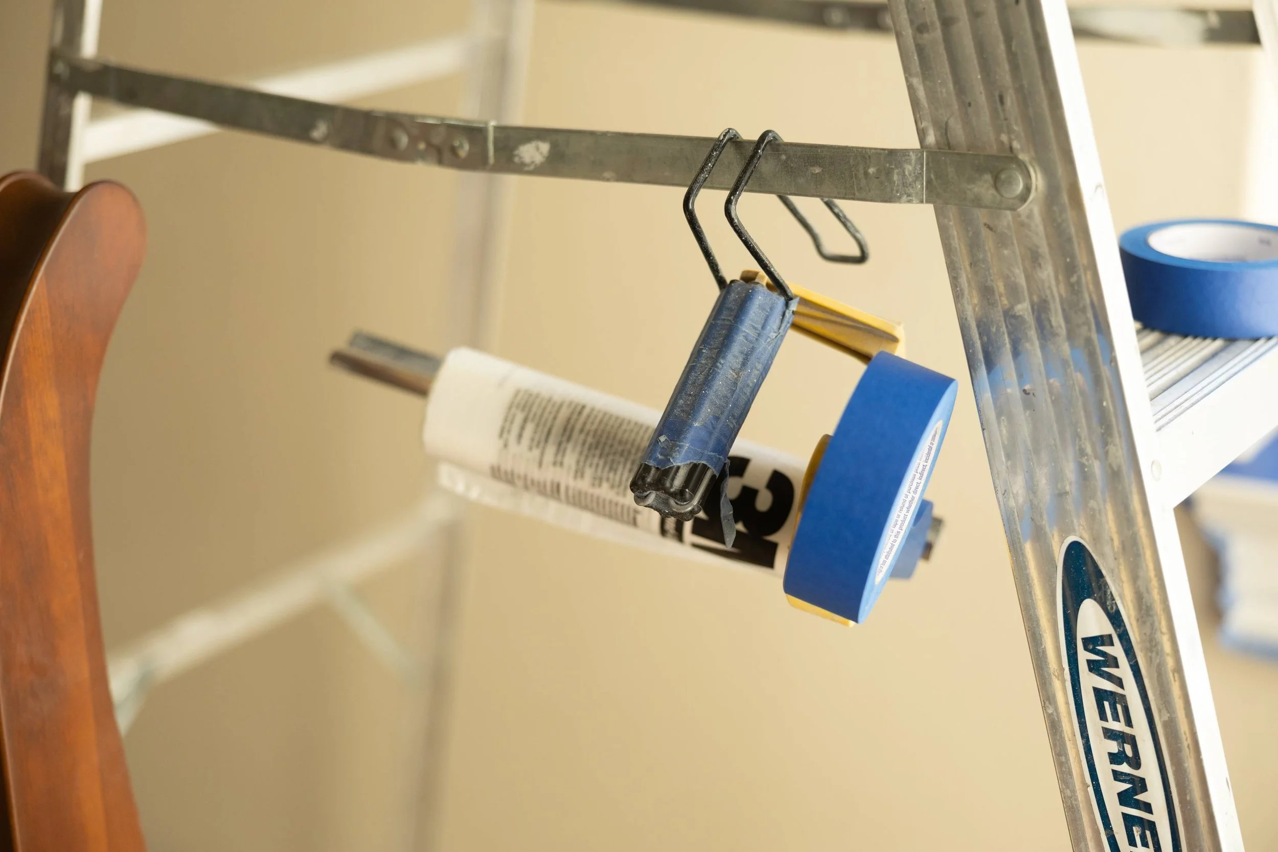Close-up of a metal ladder with painter's tape and a caulking gun hanging from it, with a partially visible wooden chair on the left.