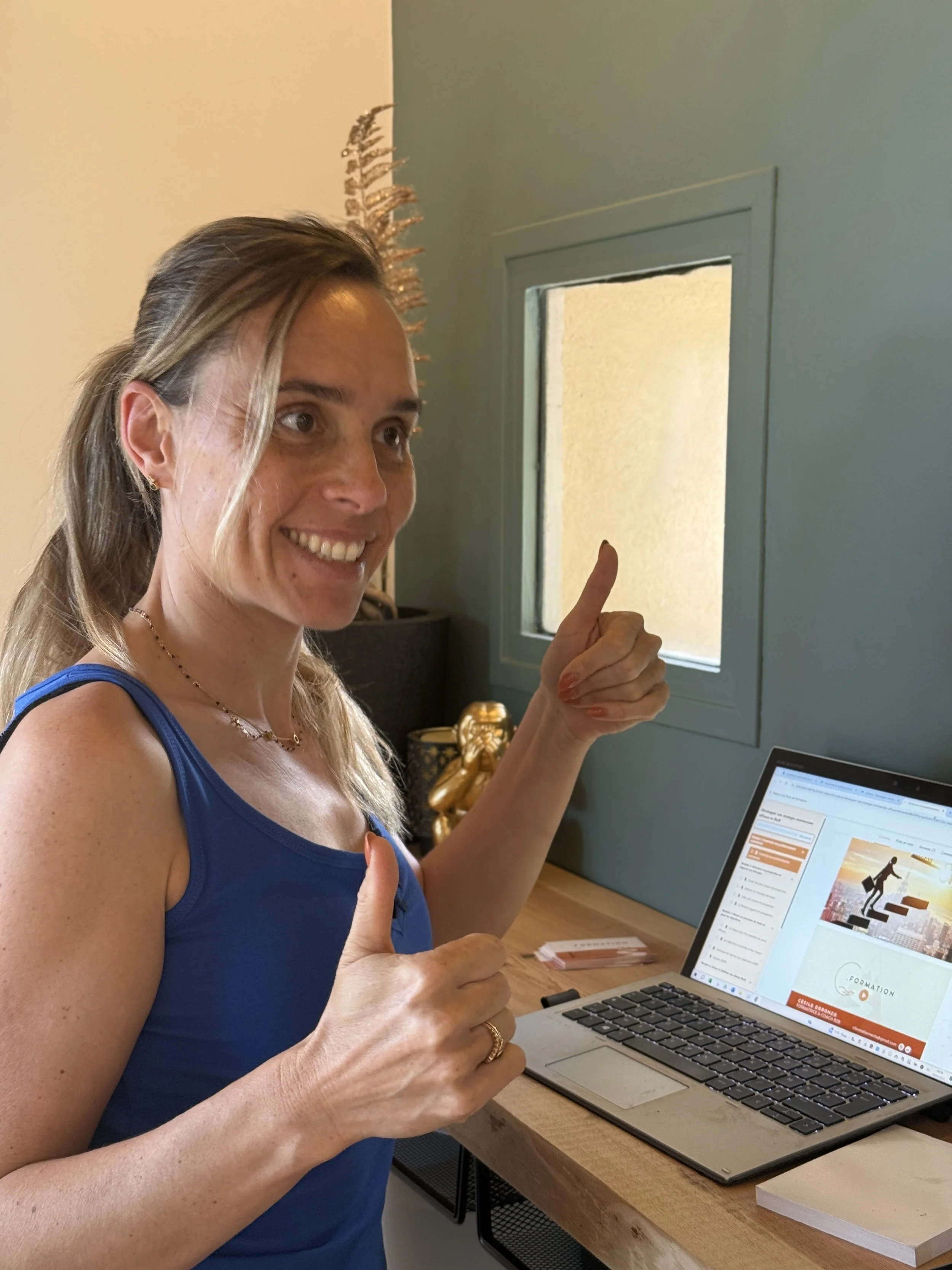 Une femme souriante en débardeur bleu, levant le pouce avec une autre main, devant un ordinateur portable, dans une pièce bien éclairée avec un mur bleu et une petite fenêtre.