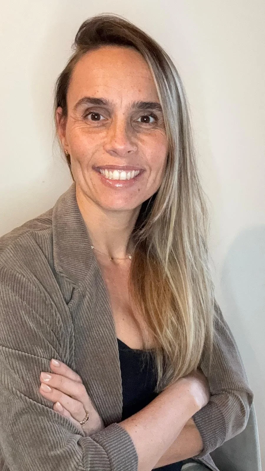 Une femme souriante aux cheveux longs et blonds, portant une veste en tissu côtelé de couleur marron, avec un fond blanc.