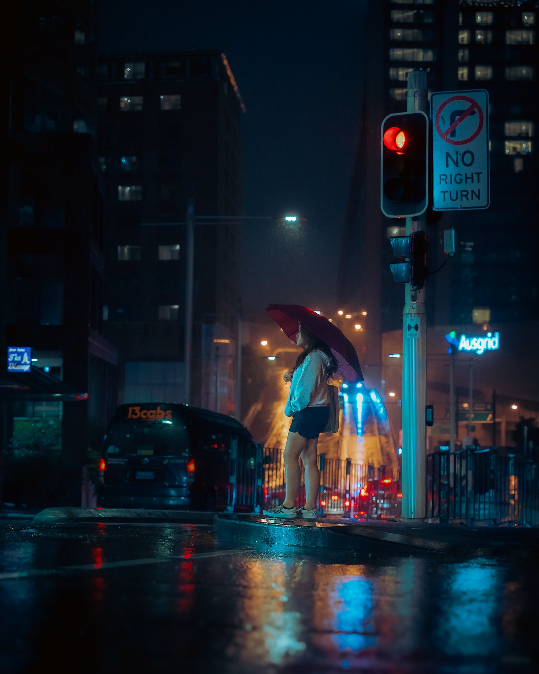 A woman holding an umbrella standing at a pedestrian crossing in the rain at night, with a red traffic light and a 'No Right Turn' sign nearby, urban background with buildings and streetlights.