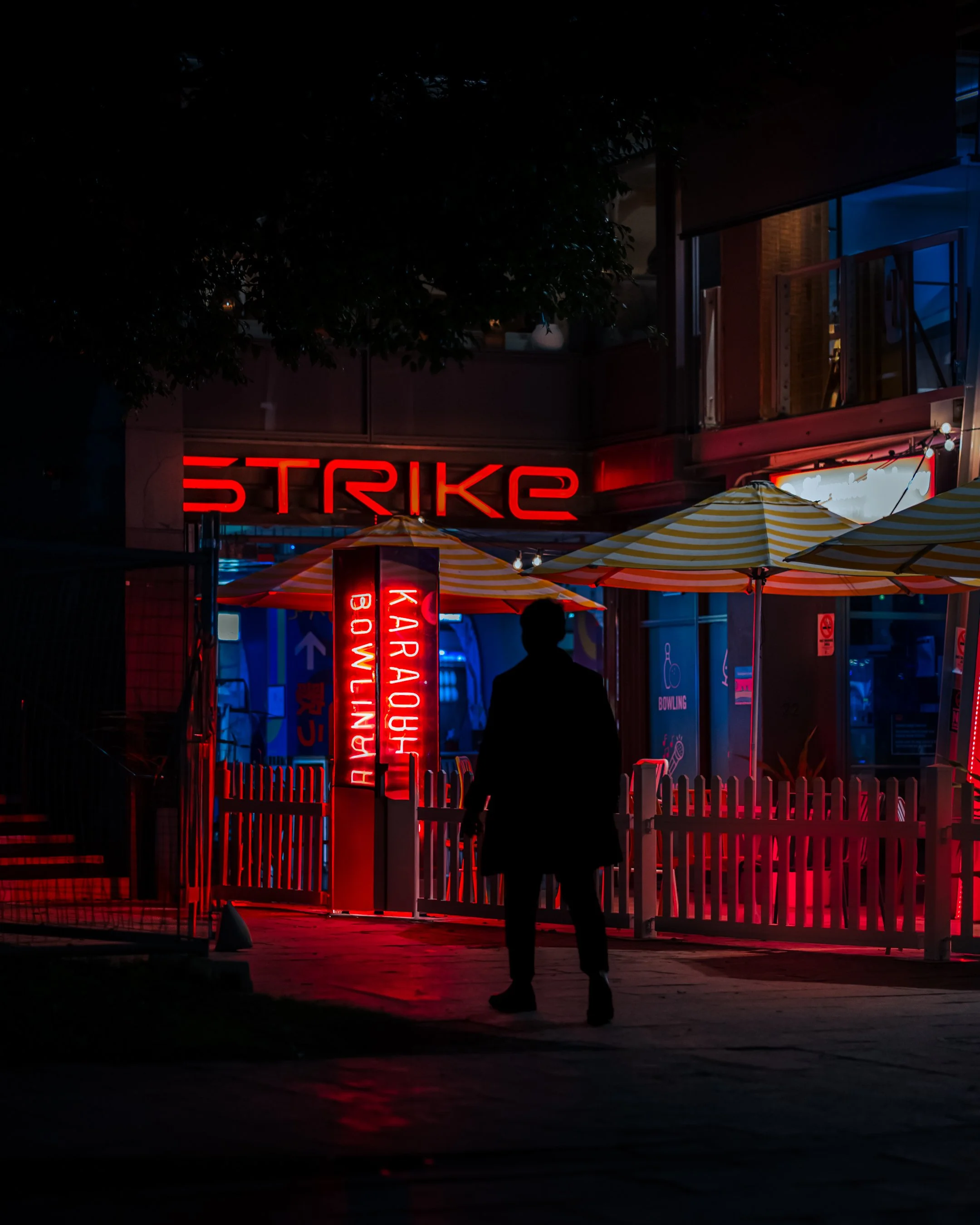 A silhouette of a person standing in front of a neon-lit establishment with red and blue signs, umbrellas, and outdoor seating at night.
