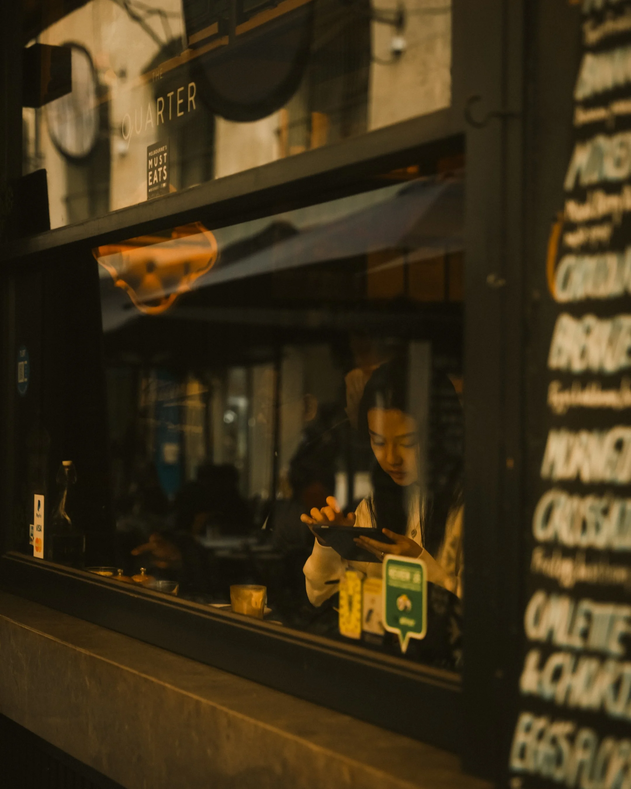 A woman inside a dimly lit restaurant or café, seen through a window, looking at a menu or tablet.