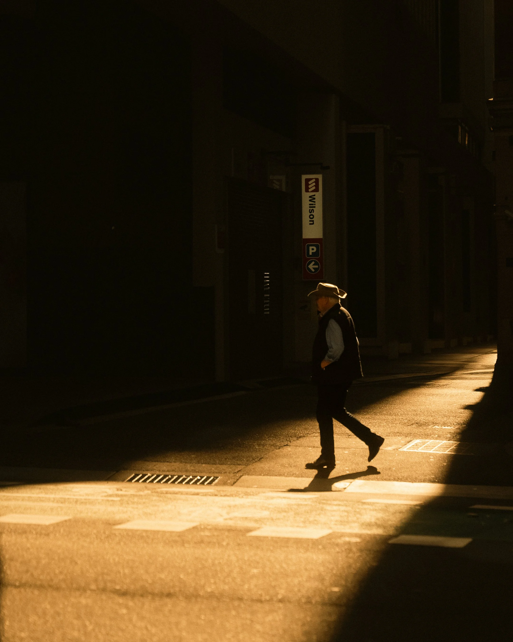 A man wearing a hat and vest walking across a crosswalk in a dimly lit street at sunset.