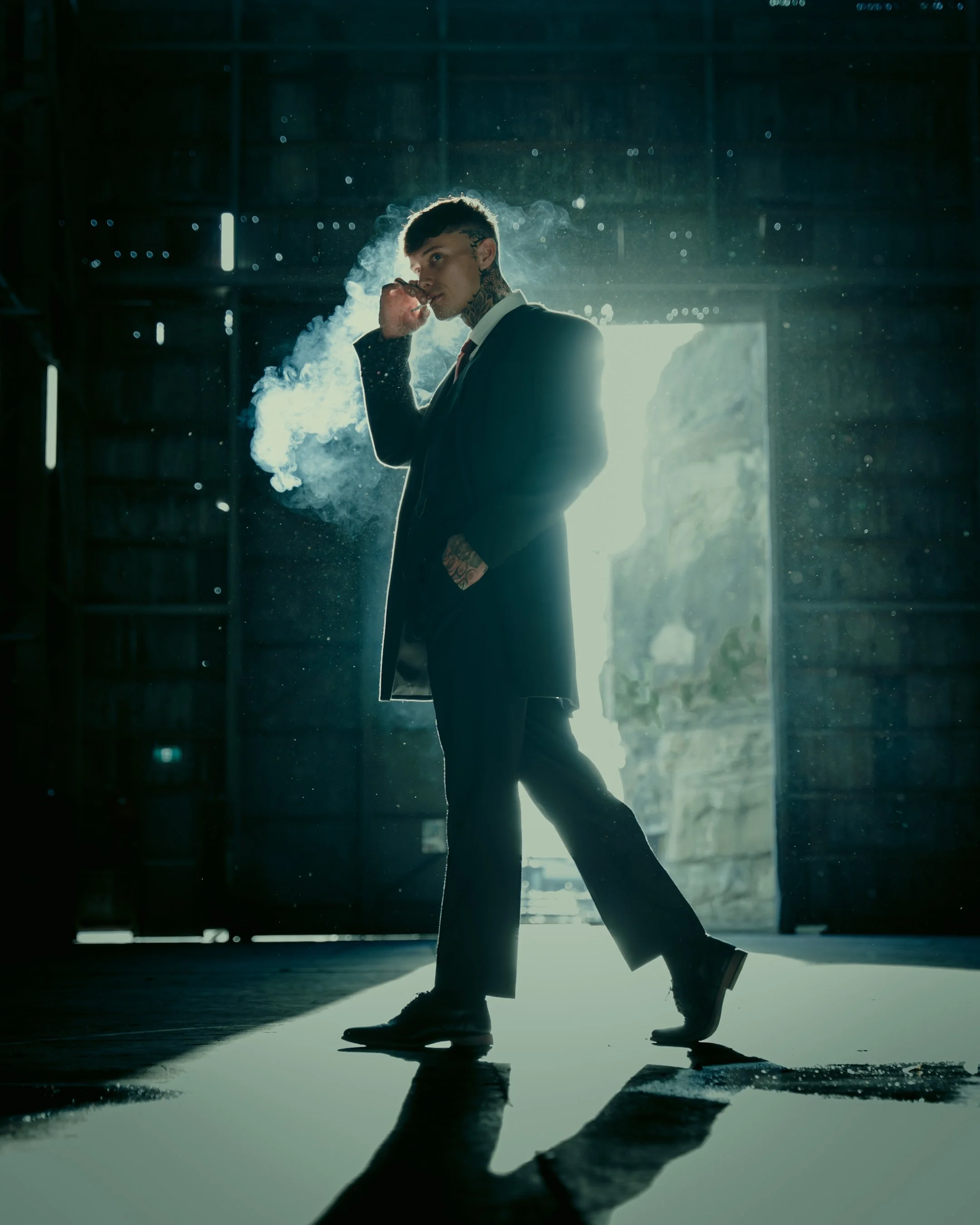 A man in a suit smoking a cigarette in a dark, industrial-style setting with light coming from an open door behind him.