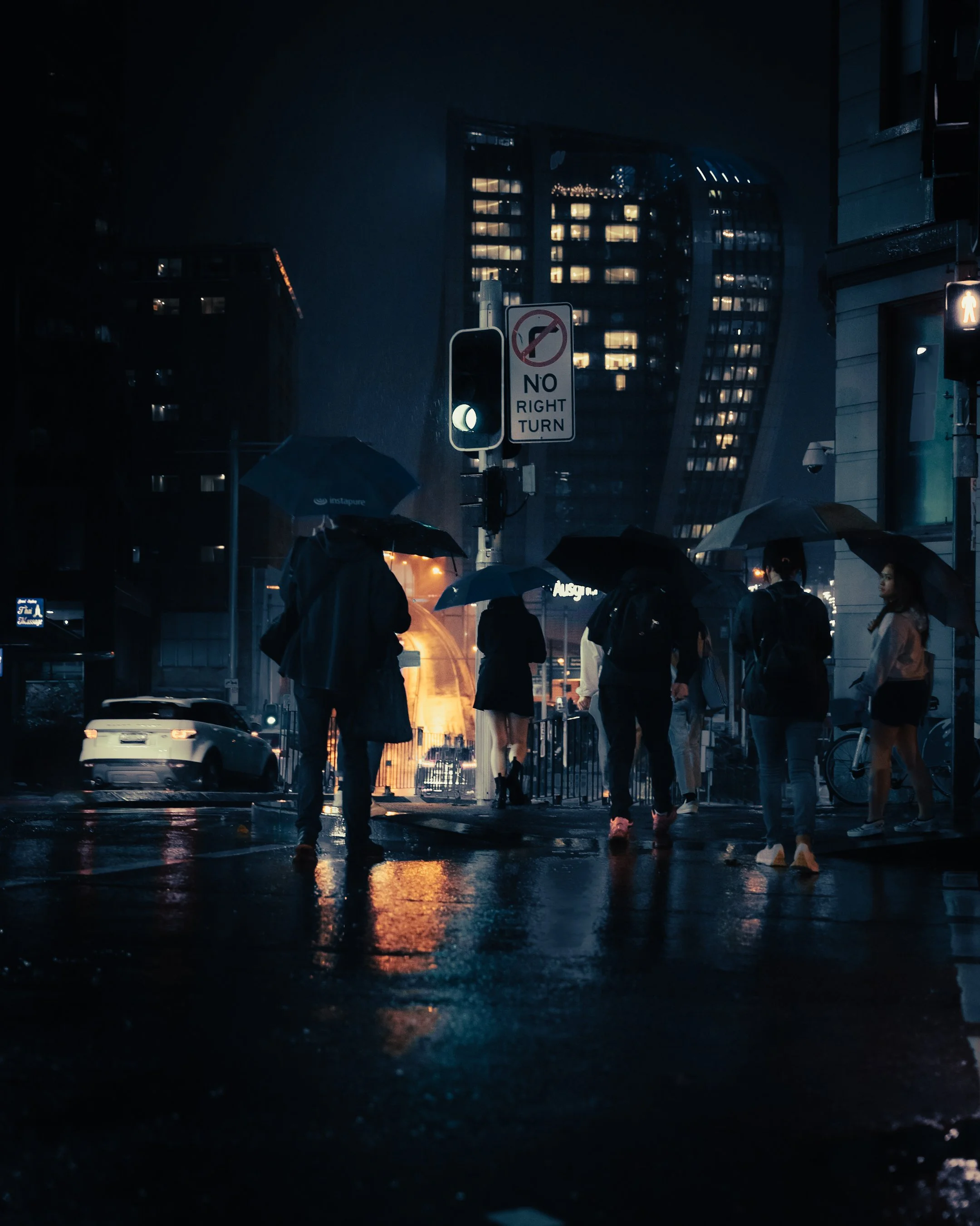 People crossing a city street at night with umbrellas in the rain, illuminated by streetlights and surrounded by tall buildings.
