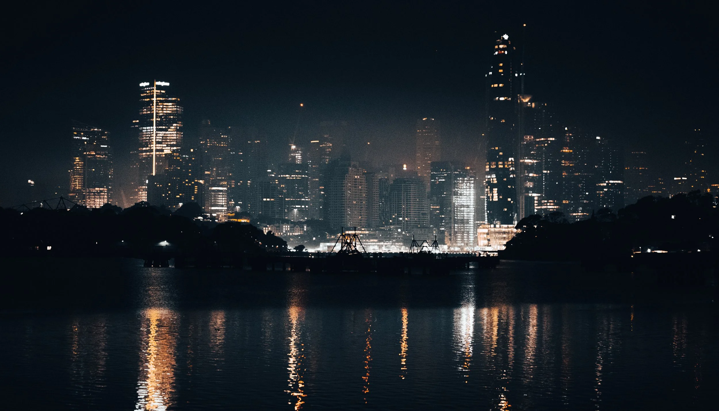 Nighttime city skyline with illuminated skyscrapers reflecting on water, dark sky in the background.