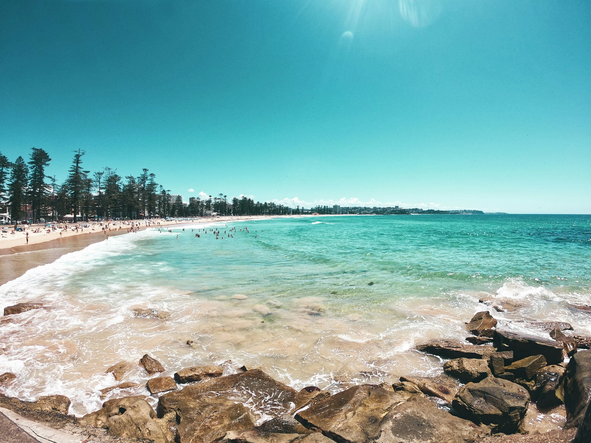A sunny beach with many people swimming and relaxing, lined with tall trees and buildings in the distance, clear turquoise water, and rocks in the foreground.