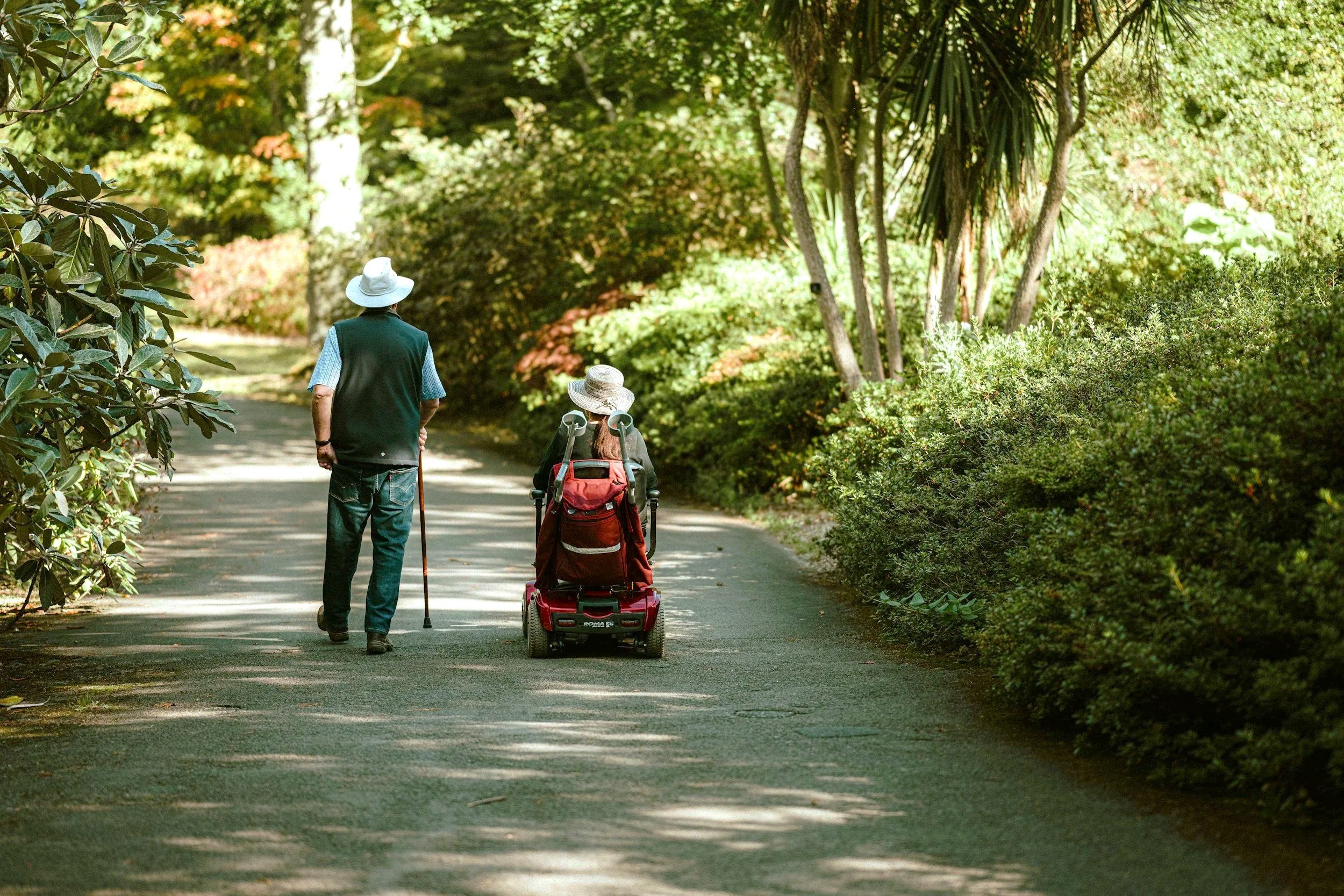 An elderly couple walking on a shaded park pathway amidst lush green trees and bushes, with the woman pushing a motorized scooter.