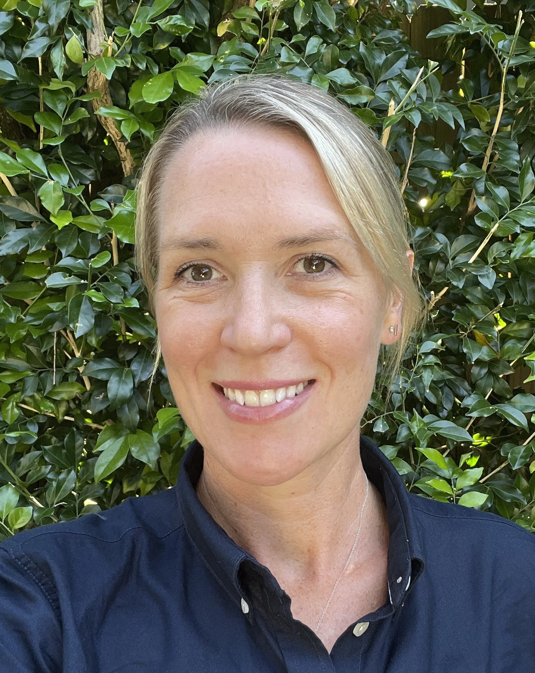 A woman with blonde hair smiling in front of green leafy bushes.