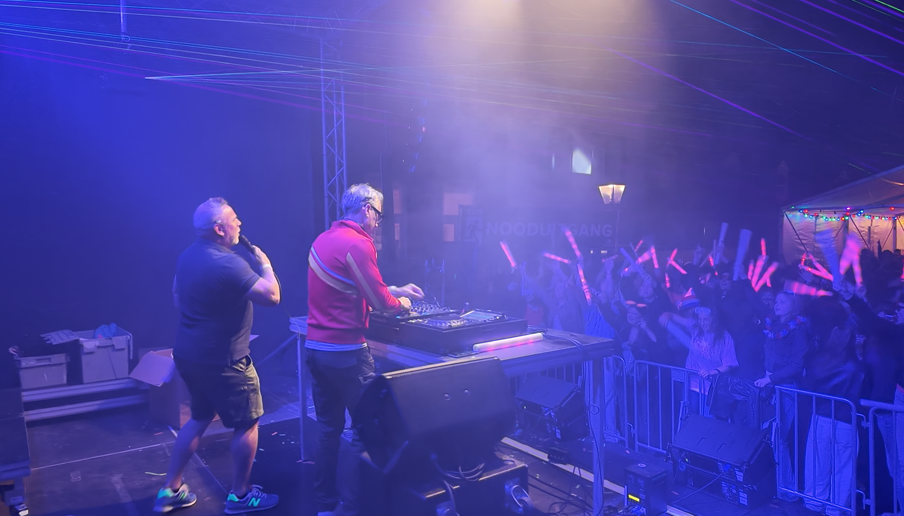 Two DJs perform on stage at a nightclub or party, with a crowd dancing and waving glow sticks in front of them, under purple and blue lighting.