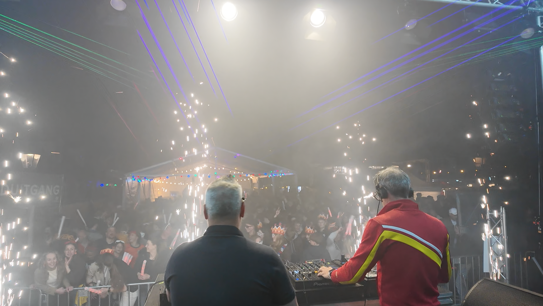 A lively indoor party or concert with a DJ playing music on a console, a crowd of laughing and cheering people, colorful lights, and sparklers creating a festive atmosphere.