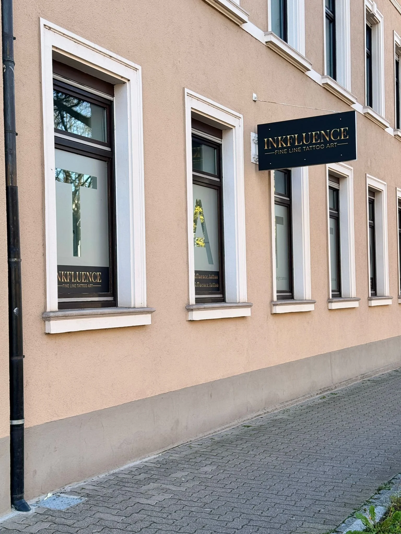 Außenansicht eines Gebäudes mit mehreren Fenstern und einem schwarzen Schild mit goldener Schrift, auf dem steht 'INKFLUENCE - FINE LINE TATTOO ART'.