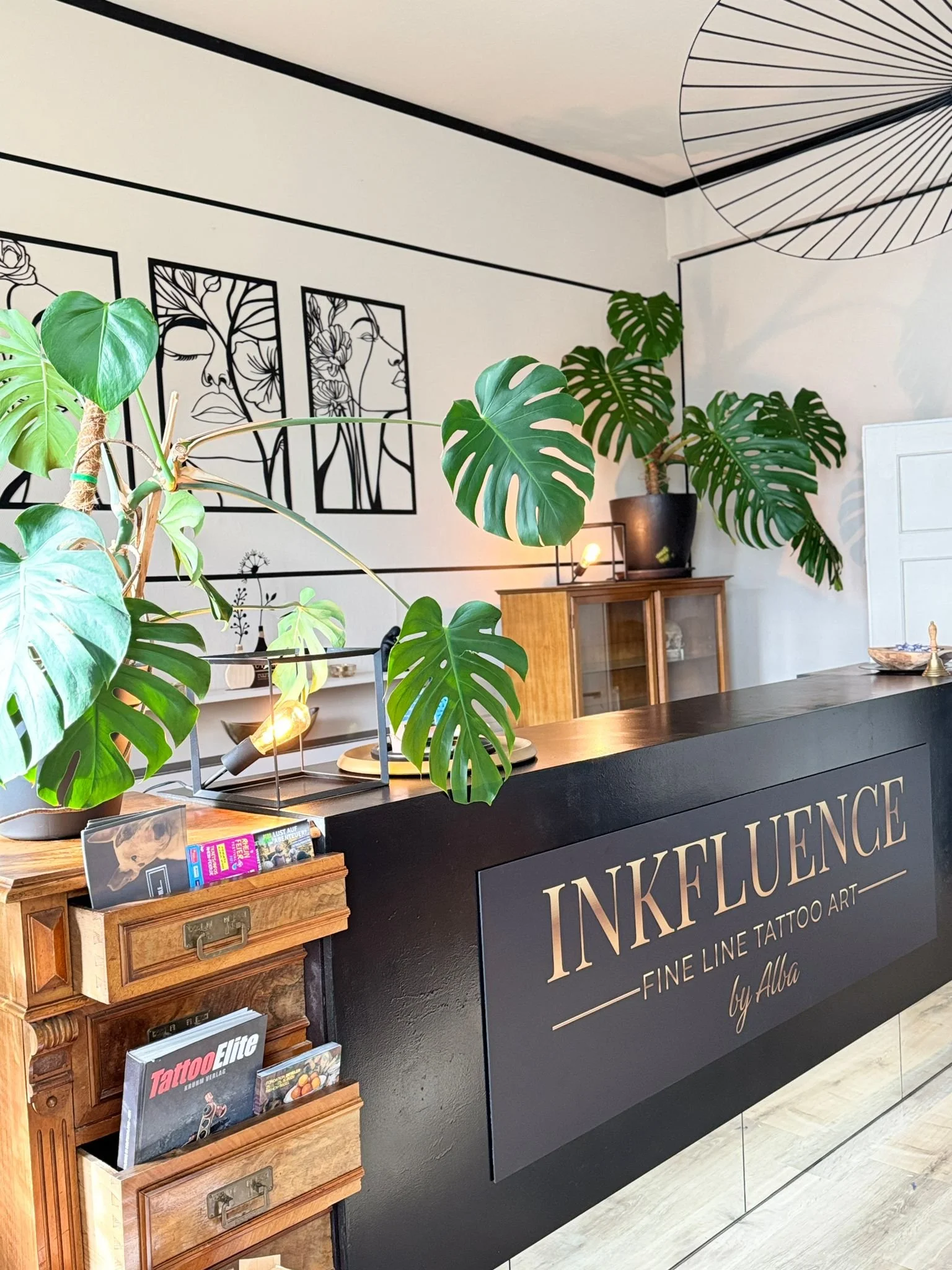 Eingangsbereich eines Tattoosalons mit schwarzen Empfangstresen, Pflanzen, Holzvitrine, Wandkunst und moderner Beleuchtung.