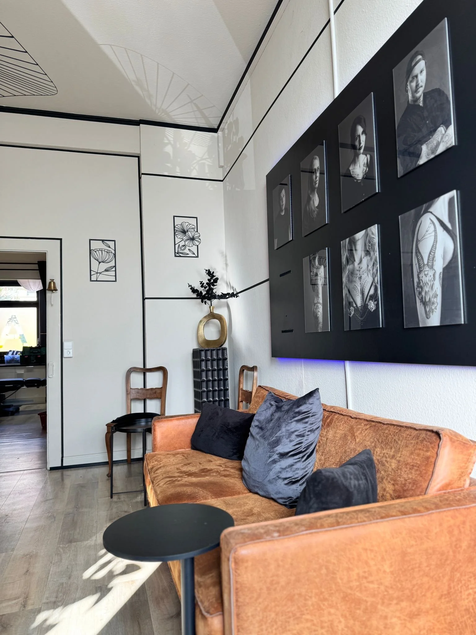 Wohnzimmer mit braunem Sofa, dunklen Kissen, schwarzem Couchtisch, Wanddeko mit Fotos und Kunstwerken, einer Stehlampe, zwei Holzstühlen, einer Vase mit Pflanzen, im Hintergrund eine Tür zu einem Raum mit Fenstern.