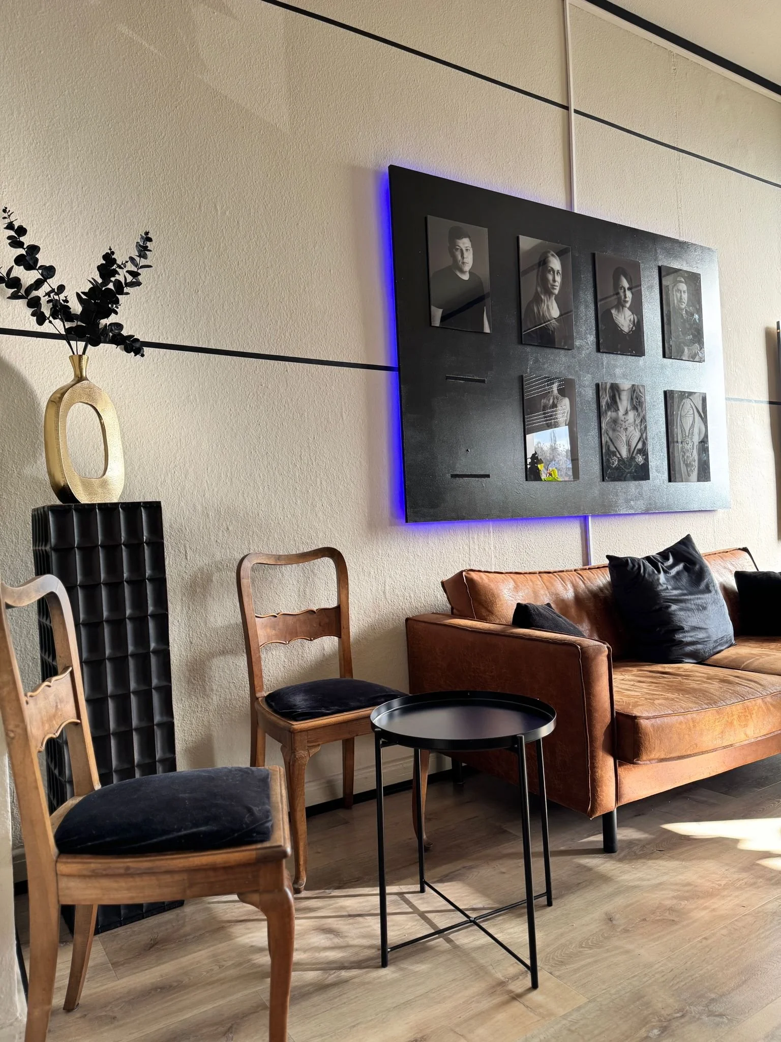 Ein Wohnzimmer mit einem braunen Ledersofa, schwarzen Kissen, zwei Holzstühlen mit schwarzen Kissen, einem schwarzen Beistelltisch, einer schwarzen Wandtafel mit Fotocollage und einer Vase mit schwarzen Zweigen auf einer schwarzen Skulptur.