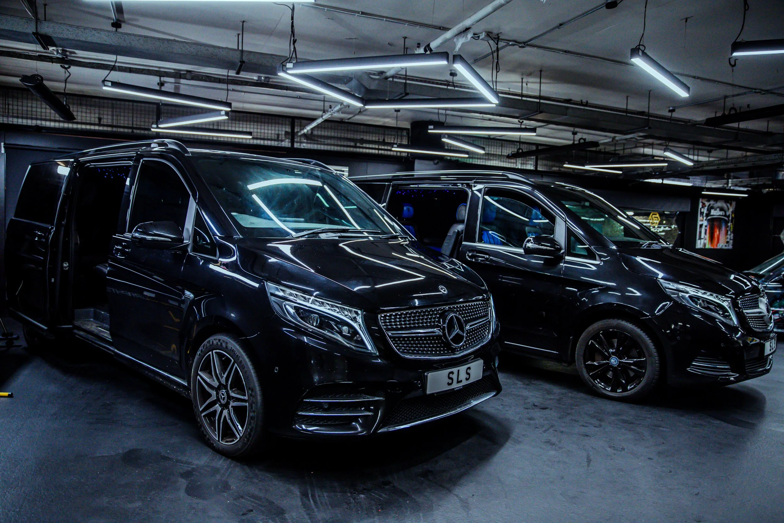 Two black luxury Mercedes-Benz vans in an underground parking garage with bright ceiling lights.