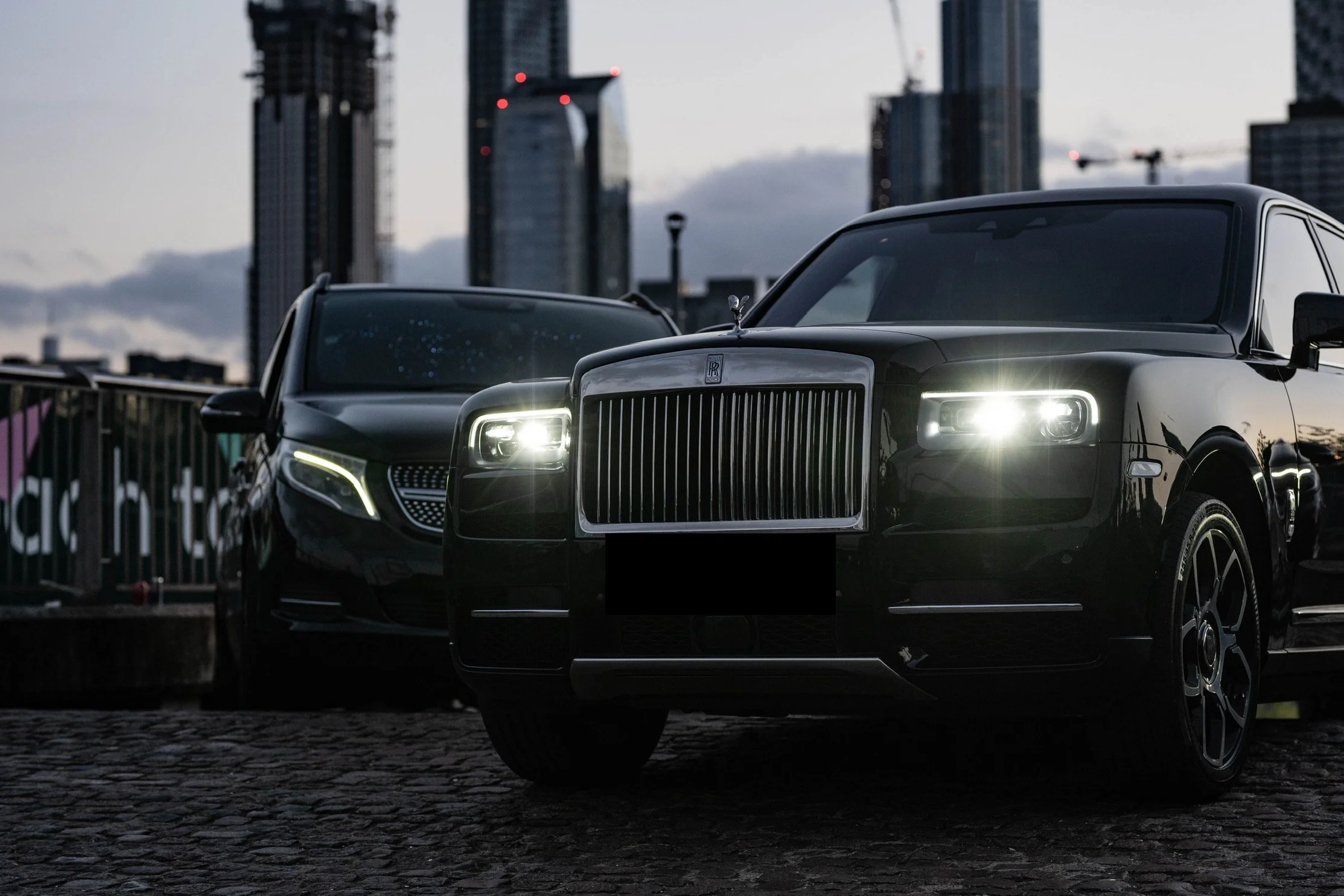 A black Rolls-Royce and a black Mercedes-Benz parked on cobblestone pavement against a city skyline at dusk.