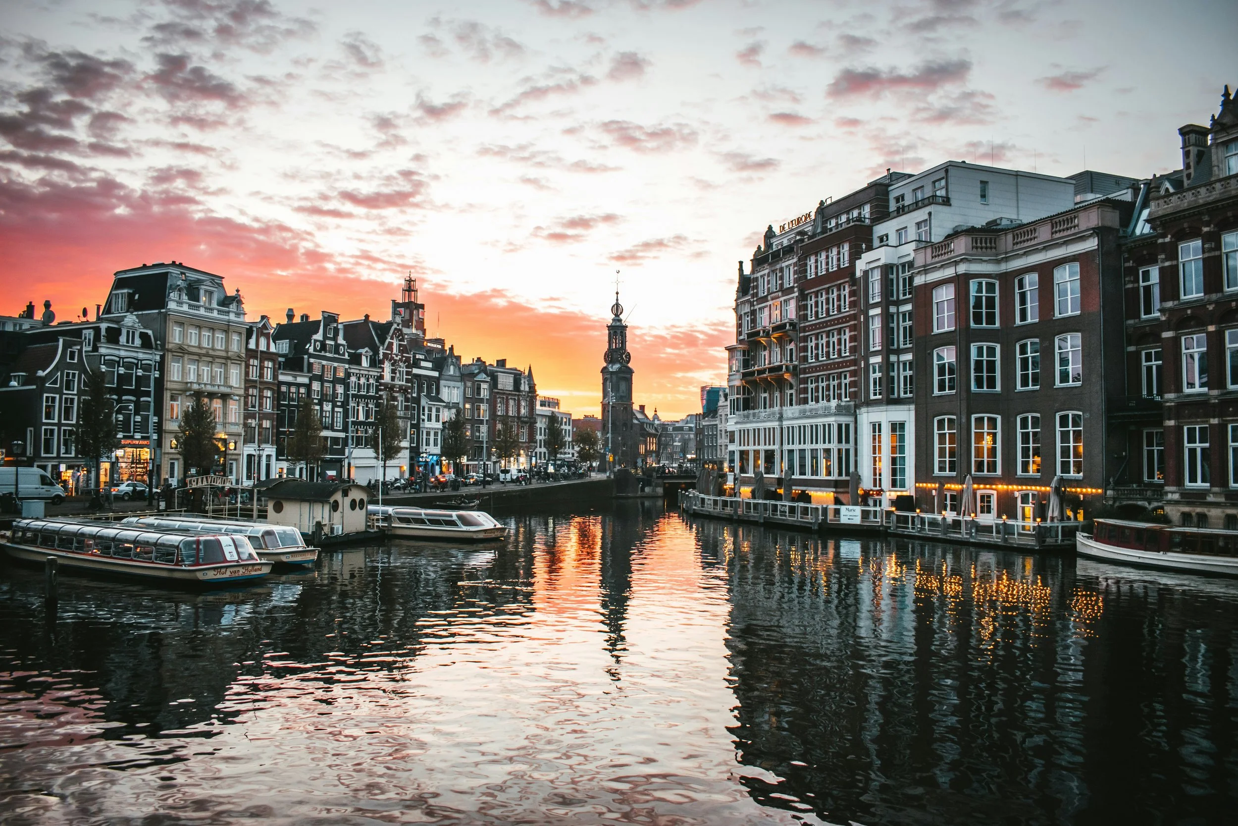 View of a canal at sunset with historic buildings along the water, boats docked, and a church tower in the distance in Amsterdam.