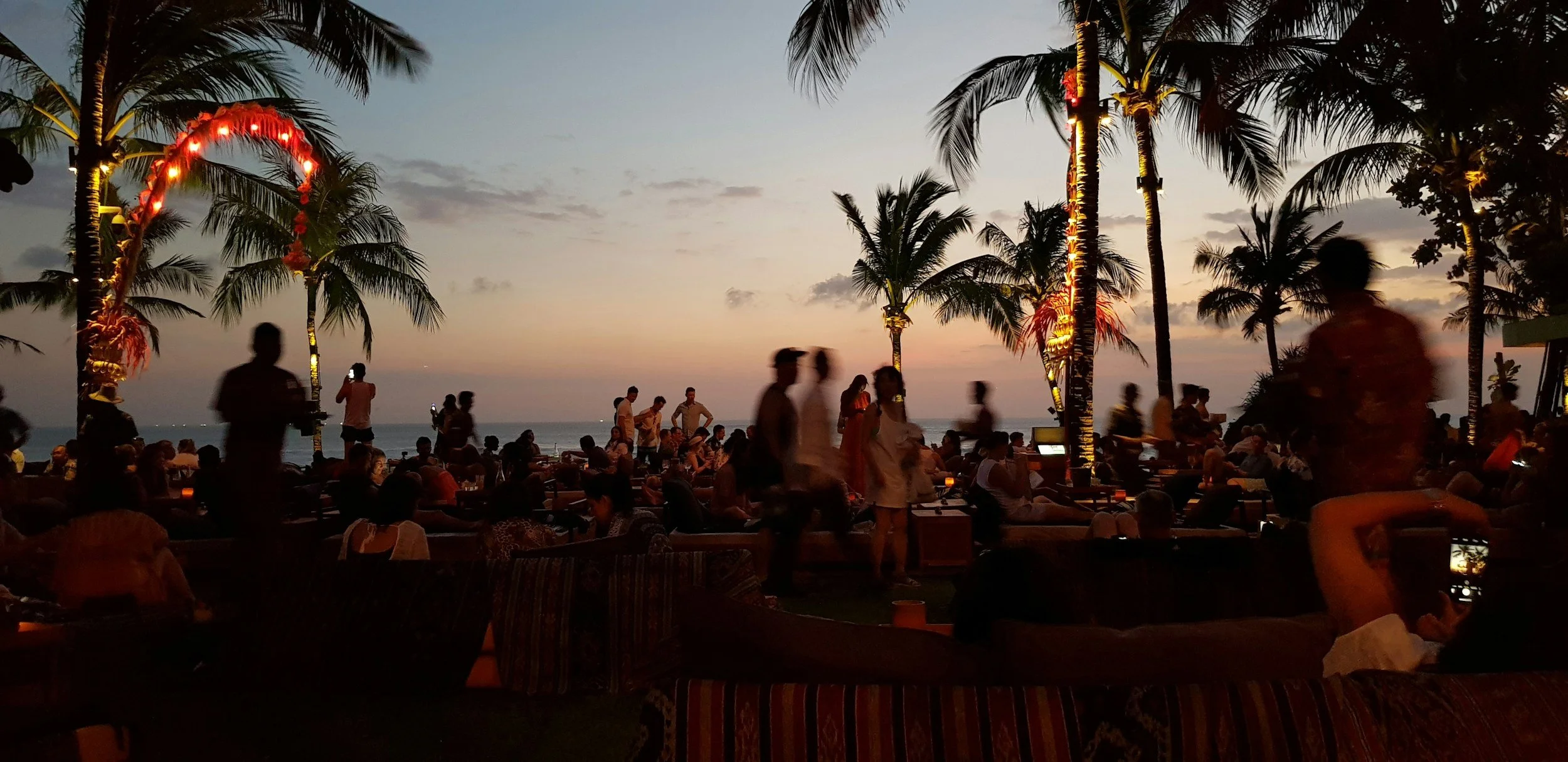 People enjoying a sunset at an outdoor beachfront venue with palm trees and decorative lighting.